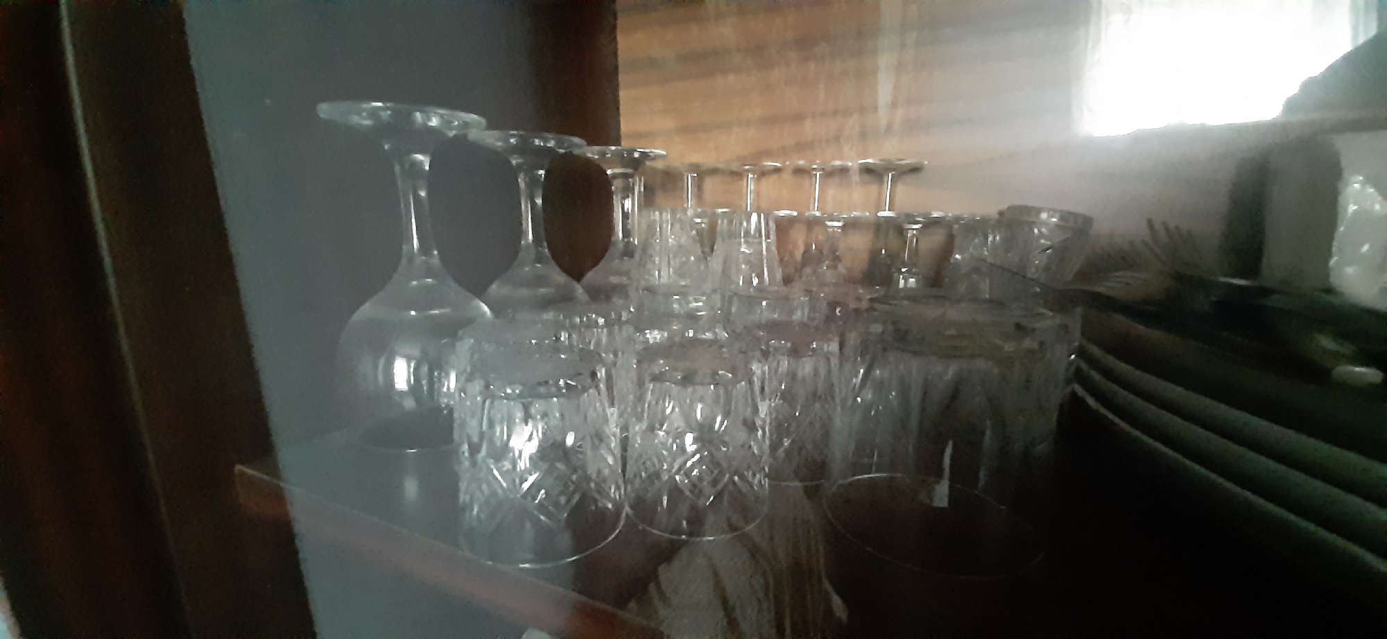 This image shows a shelf in a room, likely in Yessentukskaya, Russia, displaying a collection of glassware. On the left, several stemmed wine glasses are inverted. Behind them, a row of tumblers, some with a diamond-cut pattern, are stacked or arranged. The background appears to be a wooden cabinet or shelf unit. There are no people or animals visible in the image. The lighting is somewhat dim, with a bright light source potentially coming from the upper right, creating some glare.