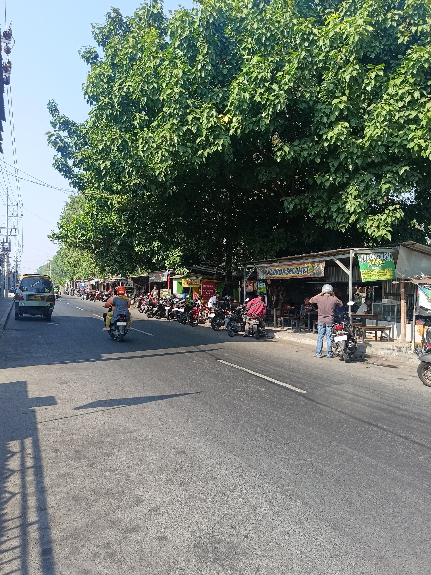 The image depicts a street scene in Calukan RT 1 RW 5 Keboansikep, Indonesia, on a sunny day with a clear blue sky. The road is paved and has a white line marking the lane. On the right side of the road, there is a row of small businesses, including a warung kopiluwak and a warung nasi. These establishments have simple structures with open fronts, tables, and chairs. Several motorcycles are parked in front of the warungs.
There are several people visible in the scene. Two individuals are riding a motorcycle in the middle of the road, with the passenger wearing a red shirt and the driver a yellow shirt and helmet. Another person, wearing a brown shirt and blue jeans, is standing next to a motorcycle, adjusting their helmet. Other individuals are seated at tables outside the warungs or walking nearby.
A green van with the text "CHYTA" and the number "35" on its side is parked on the left side of the road, facing away from the camera. Further down the road, more motorcycles and a few other vehicles are visible, suggesting moderate traffic. Large, leafy trees line the right side of the road, casting shadows on the pavement and providing shade to the businesses. Electrical poles and wires are visible on the left side of the image, indicating urban infrastructure. The overall impression is that of a typical street with local businesses and daily life in a community in Indonesia.