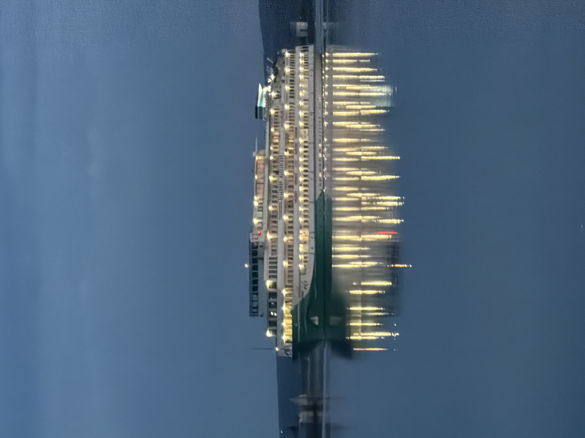 The image shows a large, lit-up ship on the water, with its lights reflecting on the water's surface. The ship is long and multi-storied, with numerous windows and what appears to be a green stripe along its side. The sky and water appear to be a dark blue, indicating either night or dusk. The location is likely near Chaikovsky, Russia, as it is a city situated on the Kama River which is large enough to host a ship of this size. The ship appears to be stationary or moving very slowly. There are no people visible in the image.