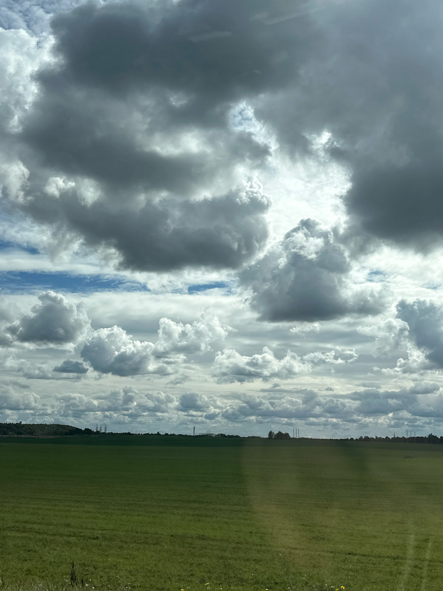 The image depicts a vast, open landscape under a dramatic sky filled with a mixture of dark, brooding storm clouds and brighter, fluffy cumulus clouds. The foreground is dominated by a large, green, cultivated field, likely a pasture or crop field, with subtle textural variations suggesting rows or growth patterns. In the distance, a treeline forms a dark, undulating horizon, punctuated by several tall, industrial structures, possibly power line pylons or chimneys, and some smaller buildings. The sky occupies the upper two-thirds of the frame, with sunlight breaking through in some areas, illuminating the edges of the clouds and casting varied light across the scene. There are no people or animals visible in the image. The location is identified as within the territory of Блонскі сельскі Савет, Belarus.