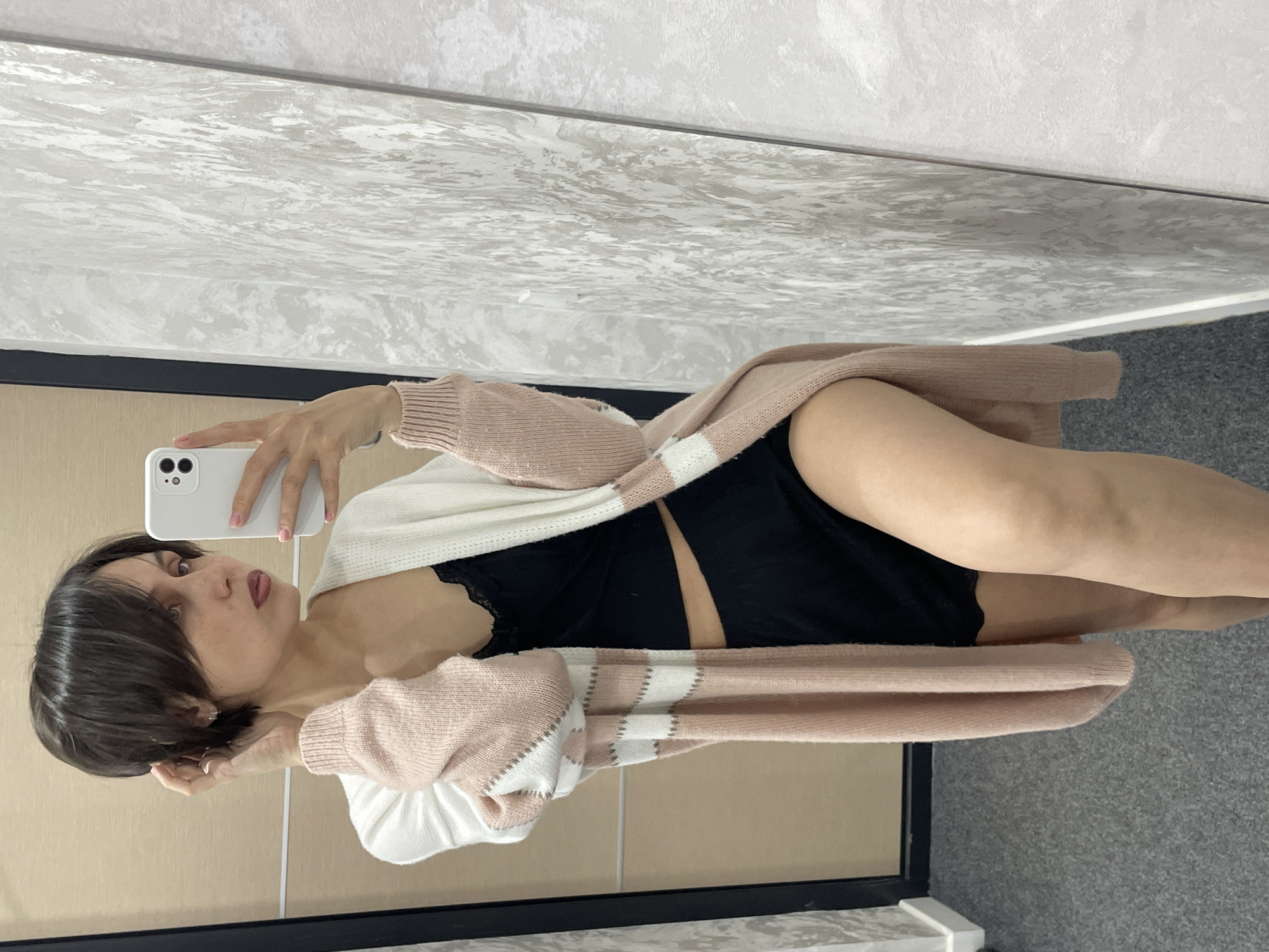 A mirror selfie captures a young woman posing indoors. She has short, dark hair, fair skin, and visible freckles on her face. Her expression is a direct gaze at the camera with a slight pout. She is holding a white smartphone, identifiable by its multi-lens camera module, in her left hand to take the photo. Her fingernails are painted a light pink.
The woman is dressed in a light pink and white horizontally striped long cardigan, which is draped over her shoulders and arms. Beneath it, she wears a black top or bralette, revealing a section of her midriff, and black shorts or underwear.
The background, visible through the mirror's reflection, consists of a wall with a textured, possibly marbled or patterned, surface in grey and white hues at the top. Below a thin black horizontal line, the wall transitions to a plainer, lighter shade, possibly a light wood panel or painted surface. The floor beneath her is covered with a grey carpet. The image is oriented sideways, appearing rotated 90 degrees counter-clockwise. The scene is an interior space, likely in Izhevsk, Russia. There is no visible text in the image.