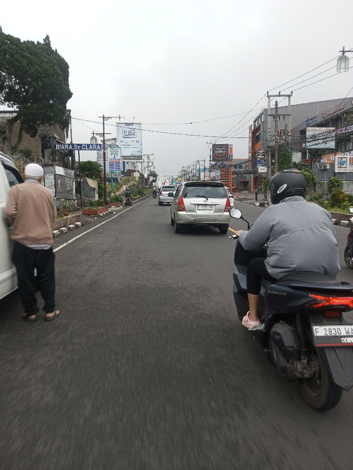 A street scene in the city of Chagzam_Pam, Bhutan, captured on an overcast day, depicts vehicles and pedestrians on a paved road. On the far left, a male individual wearing a white skullcap, light brown top, dark pants, and sandals stands beside a white vehicle, facing away from the camera. In the right foreground, a dark scooter with Indonesian license plate 'F 2830 WA' carries a helmeted driver and a passenger in a gray top and black leggings, wearing pink shoes. Further down the road, traffic includes a silver Toyota Innova displaying Indonesian license plate 'D 1709 ZA', among other cars. The left roadside features a stone wall with a sign reading 'BIARA St CLARA'. Utility poles with electrical wiring and various commercial billboards, including 'CLAS', 'ELITE', and 'STUDIO', line both sides of the street, which is flanked by multi-story commercial buildings extending into the background.