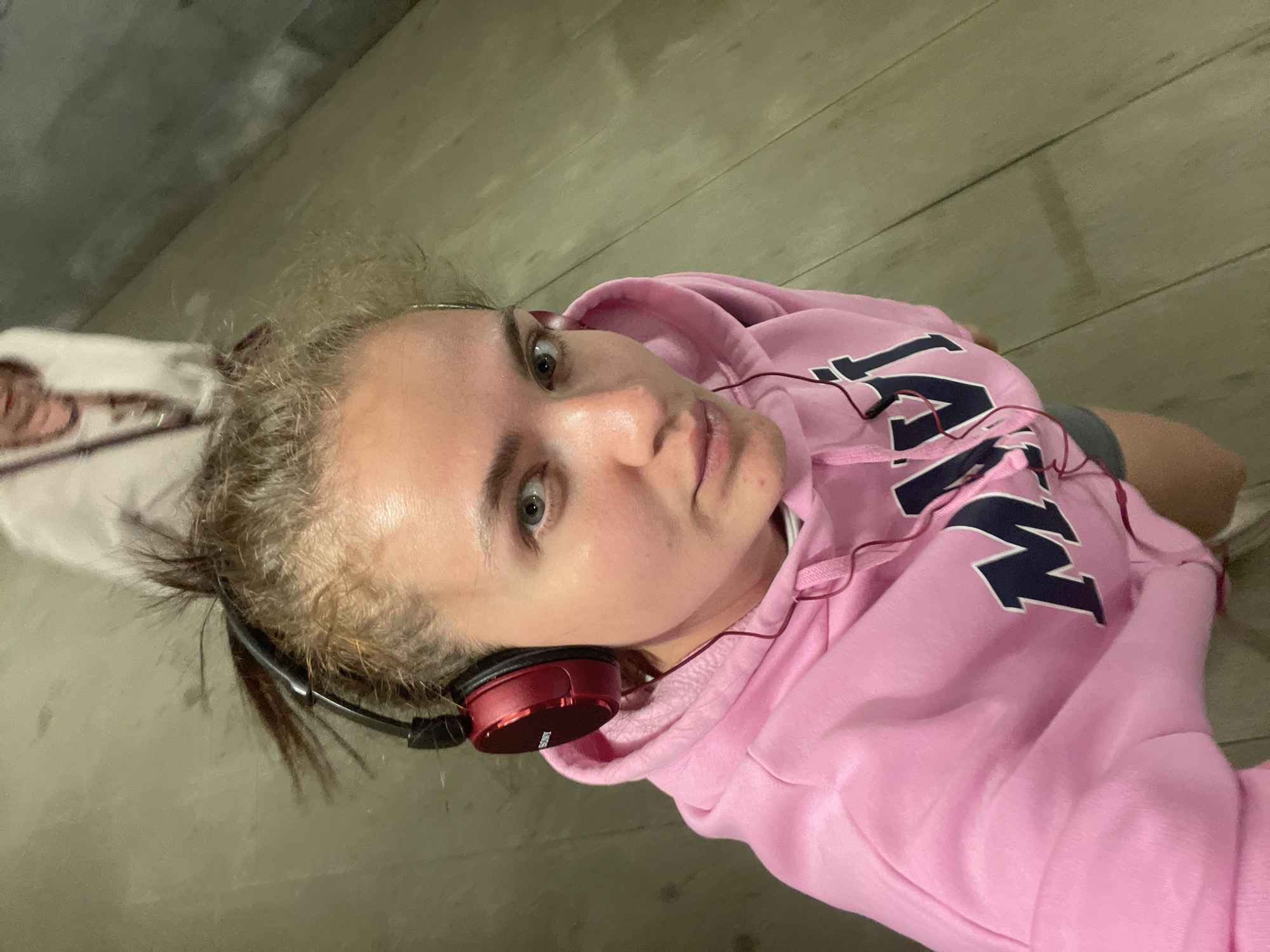 A young woman is indoors, angled upward toward the camera. She is wearing a pink hooded sweatshirt with navy blue letters and has on red and black headphones. Her hair is blonde and pulled back. The background appears to be a gray floor or wall with horizontal lines. There is no information in the image that would place the subject in Moscow, Russia.