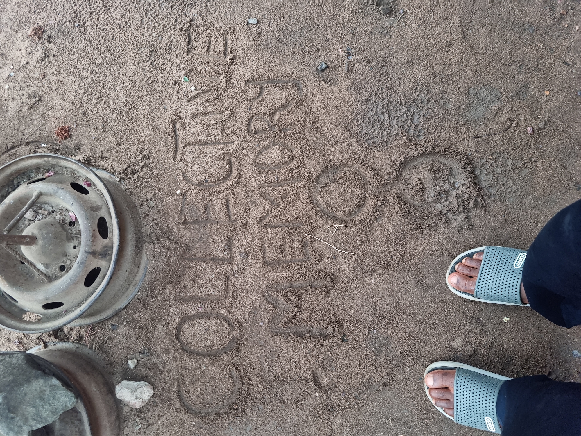 An overhead shot captures a person's feet, wearing grey slide sandals and dark trousers, standing on textured sandy-brown ground. Etched into the sand are the words "COLLEGE MEMORY" and a symbol resembling the male gender symbol (♂) next to them. To the left, an old, dirty, and metallic wheel rim rests on the ground, with a portion of another similar object visible at the bottom left. The ground is a mix of sand and small stones, suggesting an outdoor, unpaved area. The scene takes place during the daytime, likely in Fegge, Nigeria. The central activity involves someone creating sand art and then standing over it, possibly reflecting on the message.