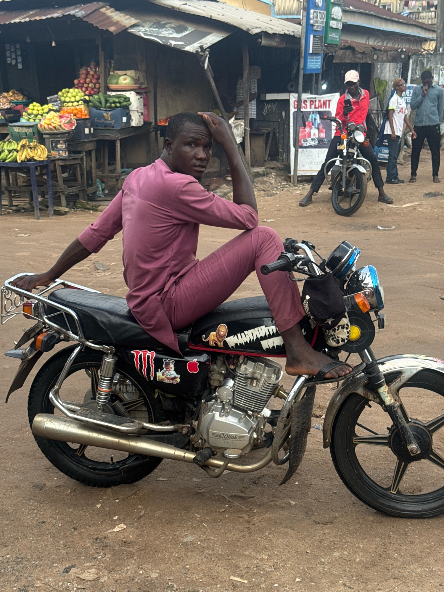 In Ihiagwa, Nigeria, a young man in a purple traditional garment sits on a black motorcycle. He rests his right hand on his head, looking towards the viewer. The motorcycle is adorned with stickers, including a lion, a monster logo, an apple, and a cartoon character. His left foot is on the footrest, and his right foot, clad in a black sandal, is on the ground. The motorcycle's exhaust pipe is prominent in the foreground. Behind him, a bustling market stall displays an assortment of fruits and vegetables. In the background, other individuals are visible, some on motorcycles and others standing, against a backdrop of market structures and buildings. The ground is a dirt surface.
