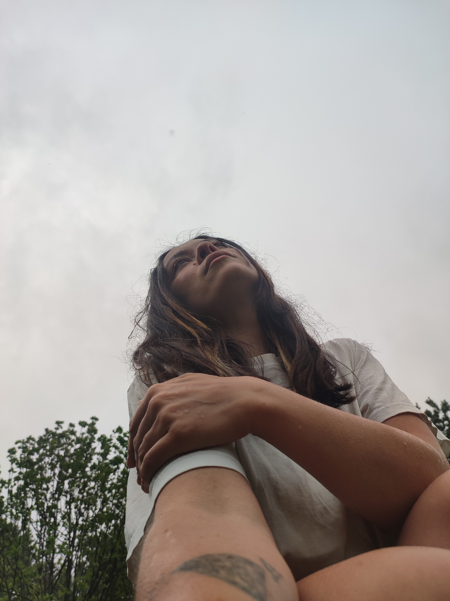 A low-angle, eye-level photograph shows a person with long brown hair, wearing a white t-shirt, looking upwards towards a cloudy sky. The person has their arms crossed. Their right arm is visible from the shoulder down, and there is a partial tattoo visible on the lower right thigh. Green trees are visible in the lower left corner of the photograph, suggesting an outdoor environment. The photograph does not provide enough information to confirm the location as Фоки, Russia, or any other specific city.