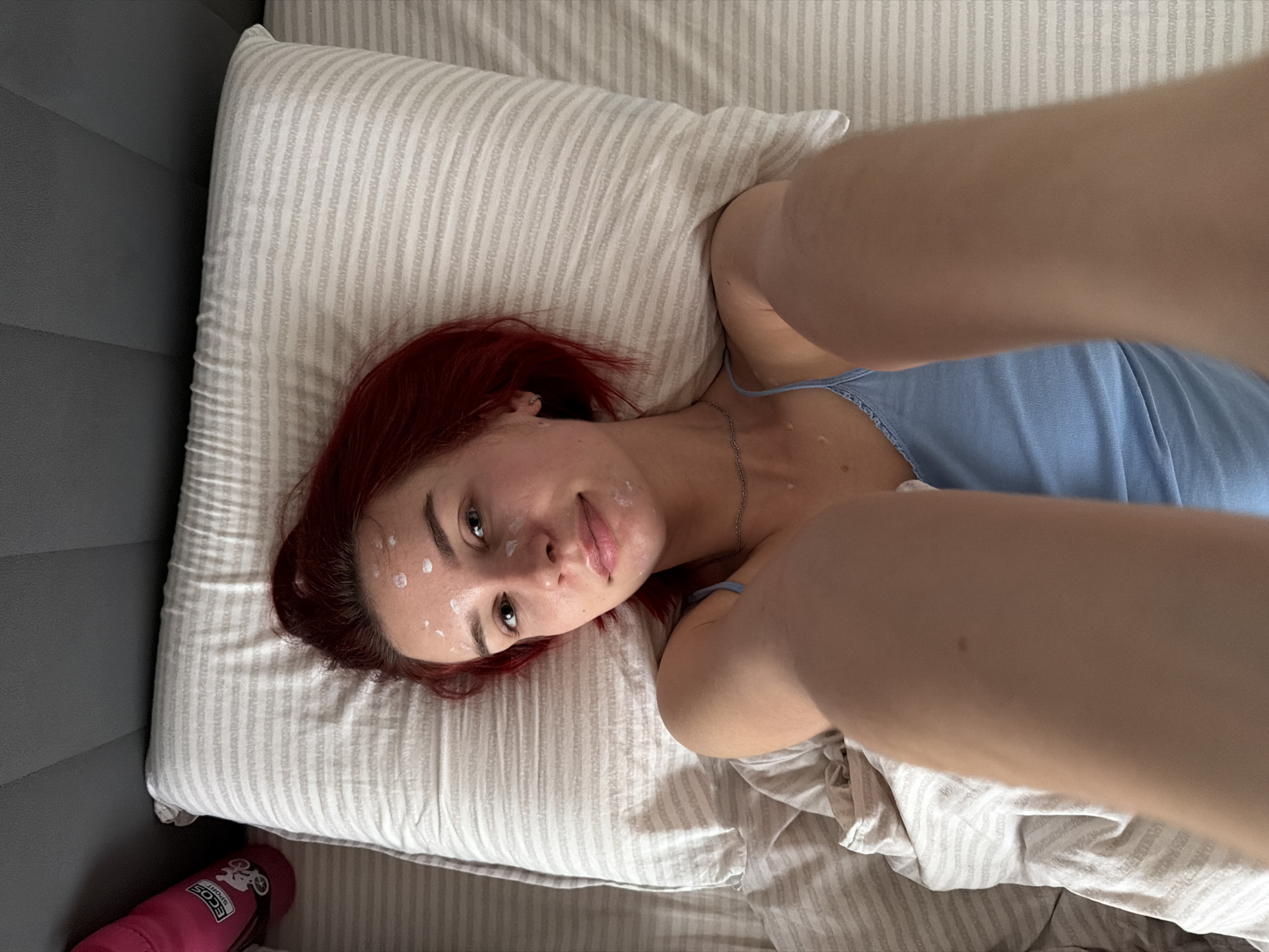 A woman with red hair is lying on a striped pillow and bedding. She is wearing a blue top and has white spots on her face. A pink water bottle is visible in the lower left corner. The scene is indoors, presumably in a bedroom. The location is Chaikovsky, Russia.