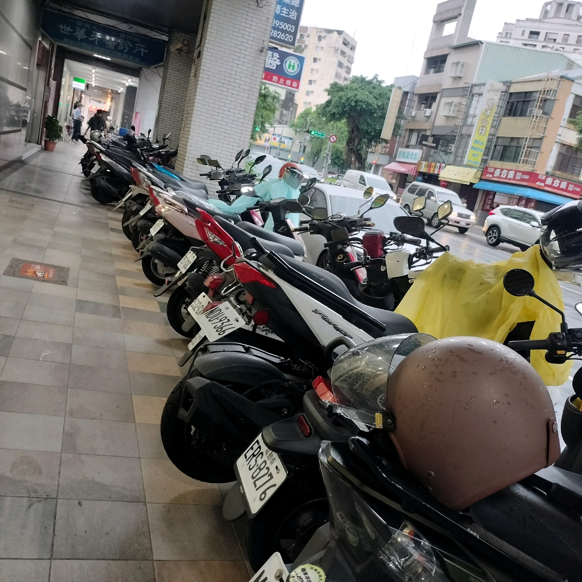 The image depicts a row of parked motorcycles and scooters on a tiled sidewalk in front of a building. The building's facade is a mix of light-colored brick and modern panels, with signage in Chinese characters. One sign appears to be for a dental clinic. Further down the street, other businesses with similar signage are visible, along with apartment buildings and trees. Several cars are also seen on the street. A person in a light blue protective suit with a face shield is partially visible in the foreground, sitting on one of the motorcycles. A brown helmet with water droplets on it is also prominent. The scene suggests a typical urban environment in Taiwan, likely Taichung, given the signage and the prevalence of scooters. The lighting and wet surfaces indicate it may be overcast or have recently rained.