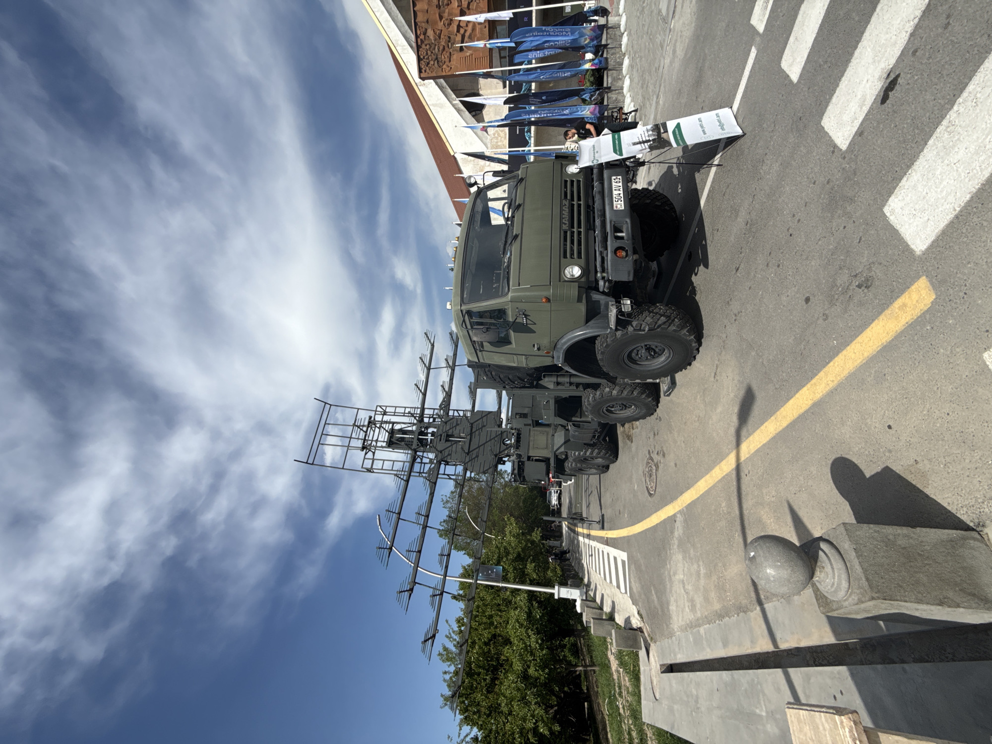 The image, originally rotated, depicts a large, olive-green military-style truck, identifiable as a KAMAZ model from the text on its grill. The truck is prominently parked outdoors on a paved road, equipped with a substantial and intricate antenna array mounted on its rear, suggesting its function as a specialized vehicle, possibly for radar or communication systems. The antenna is complex, featuring multiple rectangular and linear elements extending upwards and outwards.
The setting appears to be an exhibition or public display. In the background, there's a multi-story building with a distinctive facade and a reddish-brown roof section. Several tall, blue and white flags or banners are arranged in front of this building, displaying partial text such as "Silesian," "solutions," "silicon," and "alliance," indicating corporate or event sponsorships. A person is faintly visible behind a display stand positioned near the truck's front. This stand also shows visible text including "www.milti.com," an "Audi" logo, and "integrated solutions."
The paved road features a clear white pedestrian crosswalk (zebra crossing) and a continuous yellow line marking its edge. On the right side of the road, there's a grey concrete curb and a decorative grey stone post topped with a sphere. Trees and lush green foliage are visible in the background behind the truck. The license plate "504 AV 61" is affixed to the front of the truck.
The weather is bright and sunny, indicated by a clear blue sky with scattered wispy white clouds and distinct shadows cast by the truck, the stone post, and other elements, suggesting it is daytime, likely late morning or early afternoon. The overall impression is one of a static display of military or specialized technology at an outdoor event.