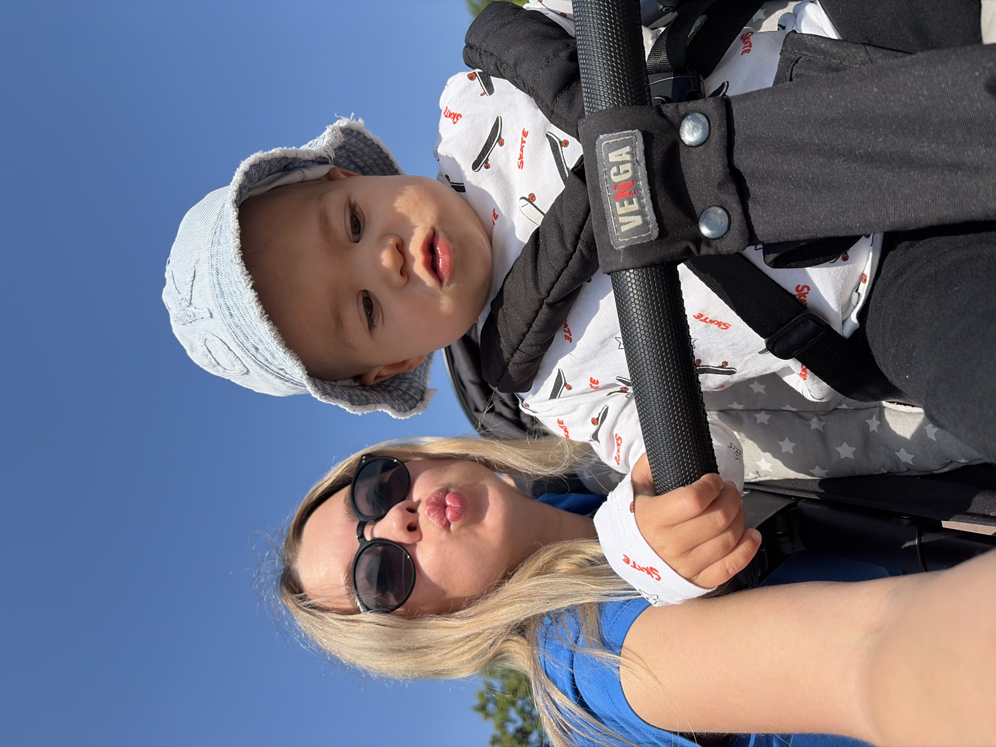 A young woman wearing sunglasses and a blue shirt is taking a selfie in a stroller with a baby. The woman is puckering her lips, while the baby, wearing a denim bucket hat and a white shirt with skateboards printed on it, looks down. The stroller has a black handle with the brand name "VEGA" visible. In the background, a clear blue sky and green trees can be seen. The image appears to be taken outdoors on a sunny day. The specific location within Chaikovsky, Russia, is not determinable from the image alone.