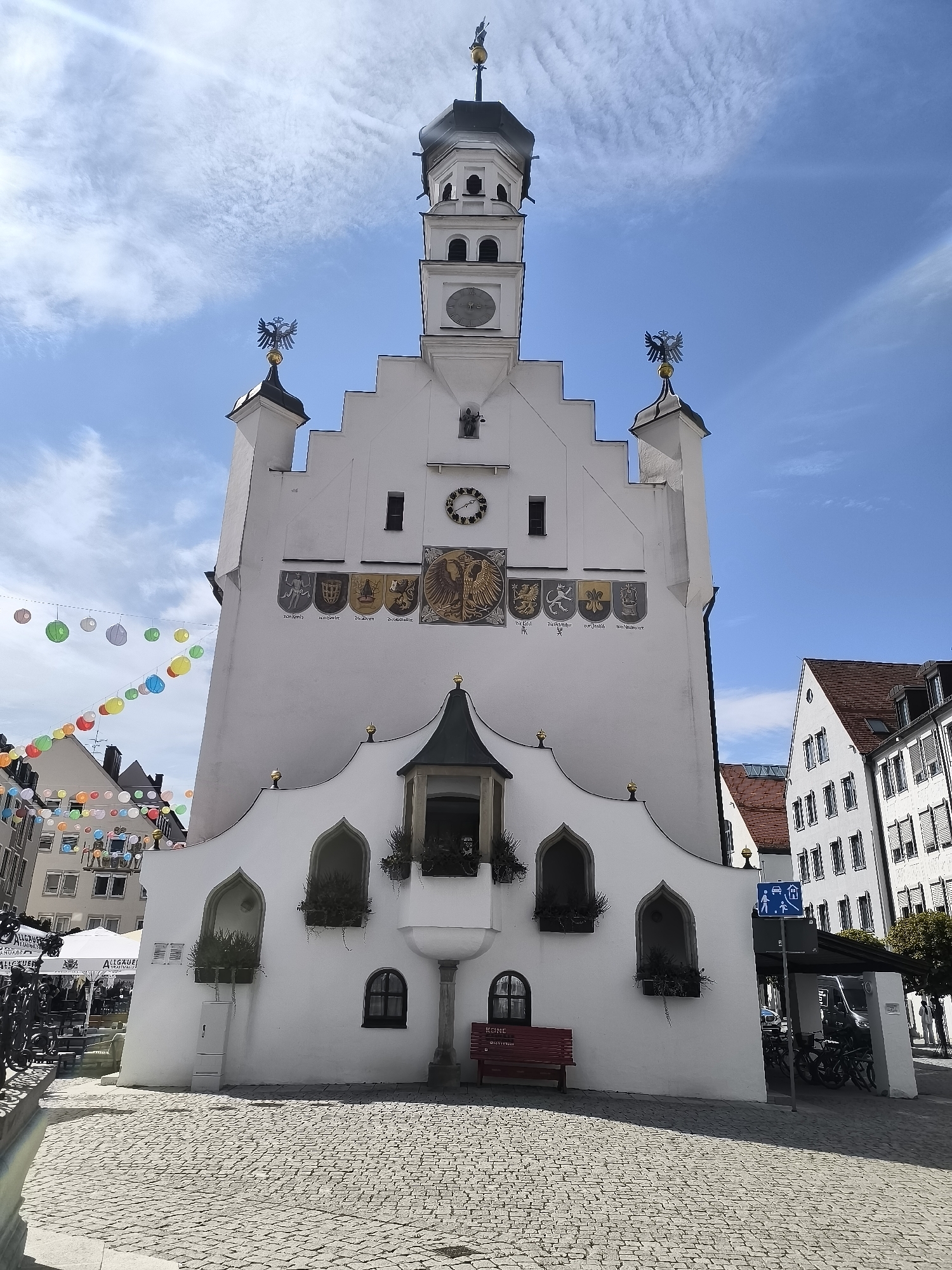 The image shows a view of the Kempten City Hall (Stadthaus) in Kempten (Allgäu), Germany. The white, multi-story building features a prominent tower with a clock face and a spire. The facade of the building is adorned with various crests and a large eagle emblem. Below the tower, a decorative section of the building has arched windows with plants and a small balcony. In front of the building, a cobblestone plaza is visible, with a red bench placed in front of the main structure. To the left, there are tents and tables set up, suggesting an outdoor market or event. Colorful paper lanterns are strung across the sky in the background. To the right of the city hall, other buildings with tiled roofs and windows are visible, along with a covered area that appears to be parking for bicycles. A blue traffic sign indicating pedestrian and vehicle access is also present. The sky is blue with scattered white clouds, and the sun is shining, casting shadows on the plaza. There are no visible people interacting with the main subject in the foreground.