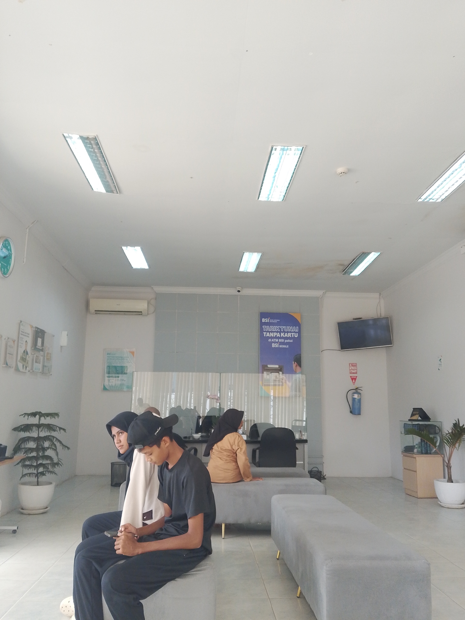 This image captures the interior of what appears to be a waiting area, likely a bank or service center, in Manetunong, Indonesia. Three individuals are visible, seated on grey upholstered benches. In the foreground, a young man wearing a black t-shirt and cap is engrossed in his phone, seated next to a young woman in a white top and black hijab, who is looking towards the left. Further back, another person in a light brown top and hijab is seated on a bench, facing away from the camera, behind a glass partition.
The setting features white walls and a light-colored tiled floor. The ceiling is white with several long, recessed fluorescent lights providing illumination, along with a visible smoke detector. A wall-mounted air conditioner is on the left wall. Various items adorn the walls: a round wall clock on the far left, several posters and flyers, and a prominent blue poster on the back wall. This blue poster clearly displays text: "BSI", "TARIK TUNAI TANPA KARTU", "di ATM BSI pakai", and "BSI mobile", indicating this is a branch of Bank Syariah Indonesia.
Other objects include a wall-mounted television screen that is off, a blue fire extinguisher below it, and a small wooden cabinet with a potted plant on the right side of the room. A large potted plant is also present on the left side near the foreground subjects. The scene suggests a calm, routine atmosphere of people waiting for a service.