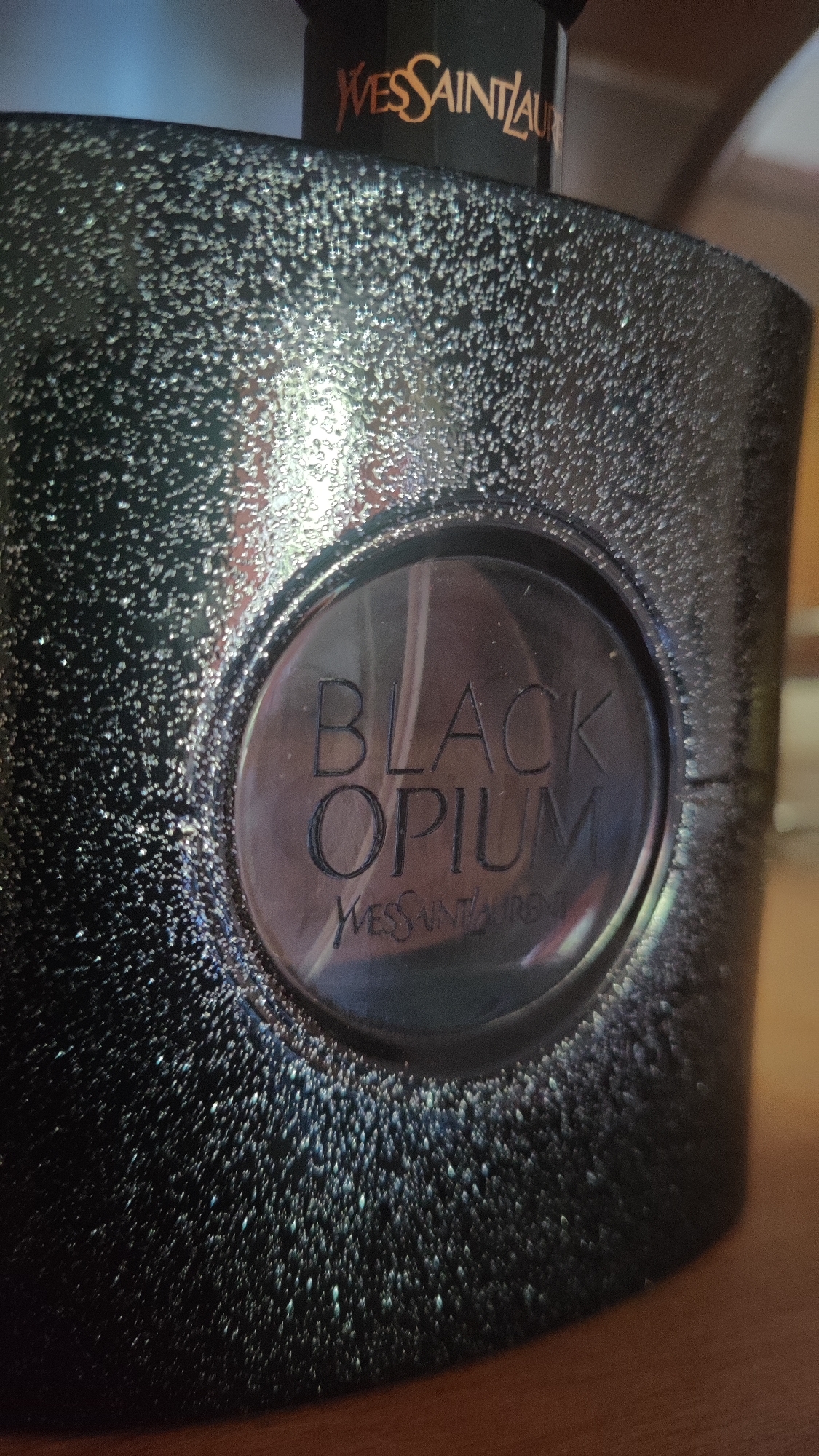 The image shows a close-up of a black, glittery perfume bottle, specifically Yves Saint Laurent's Black Opium. The bottle has a circular indentation on its front with the words "BLACK OPIUM" embossed in a darker shade. Below this, "YVES SAINT LAURENT" is also embossed. The bottle's surface is textured with numerous small, reflective specks, giving it a sparkling appearance. The top portion of the bottle, partially visible, displays the "YVES SAINT LAURENT" logo in gold lettering. The background is blurred but suggests a wooden surface and hints of a blue and brown environment. The location of this image in Dagomys, Russia, is not discernible from the visual information provided.
