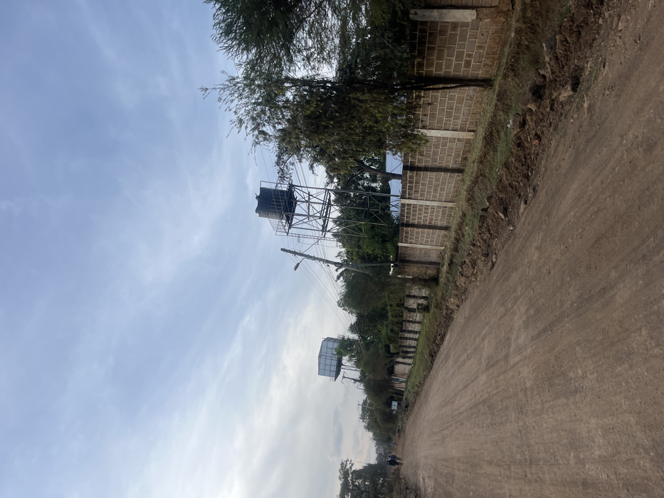 A long, unpaved road stretches into the distance under a cloudy sky. Tall trees and a block wall line the right side of the road. Water towers stand prominently on the left side, appearing twice in the frame. Power lines run parallel to the road. The scene evokes a sense of rural or semi-urban development.