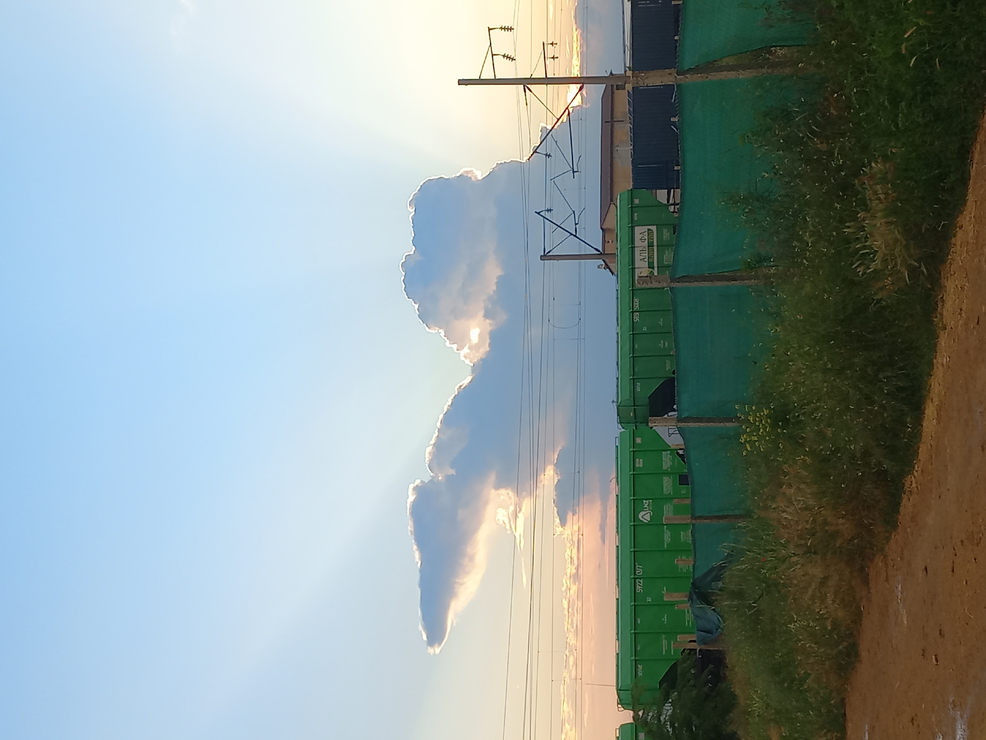 The image depicts an outdoor scene with a vast sky filled with light blue hues, featuring crepuscular rays emanating from behind a large, illuminated cloud. These rays are visible as bright beams of light stretching across the sky. Below the sky, there are several elements that suggest an industrial or transportation hub. Prominently, a line of green freight train cars stretches horizontally across the mid-ground. These cars have visible markings and numbers, such as "5722 1317" and "5929 5008." A green mesh fence runs in front of the train cars, and beyond them, a building with a brown roof is visible. Electrical wires and utility poles are also present, indicating a network of infrastructure. The foreground consists of a dirt path and overgrown green grass, suggesting an undeveloped or transitional area. The lighting and cloud formation suggest it is either sunrise or sunset. The scene is identified as being located in the city of Molodizhne, Ukraine.