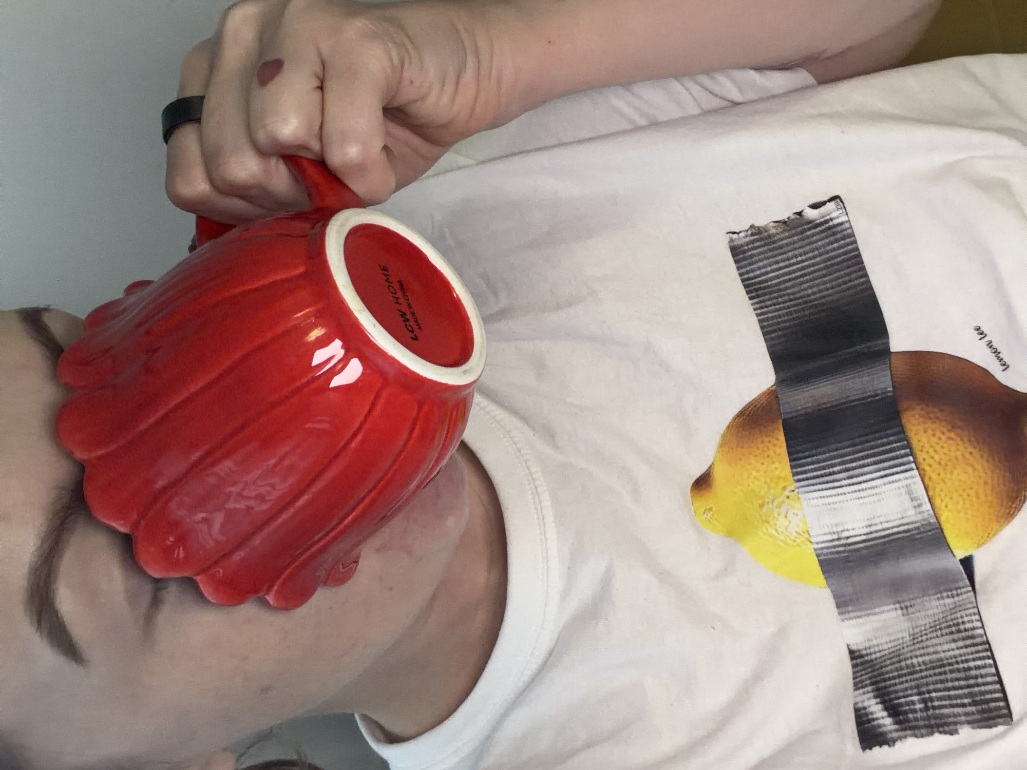 A close-up, slightly angled overhead shot depicts a person lying down, holding a large, red, fluted mug to their face. The mug's base is visible, revealing "LCW HOME" printed inside. The person is wearing a white t-shirt featuring a graphic of a lemon with gray tape across it. A hand wearing a black ring is visible holding the mug. No specific details suggest a location in Moscow, Russia, or any other specific location. The focus is on the person, the mug, and the t-shirt.