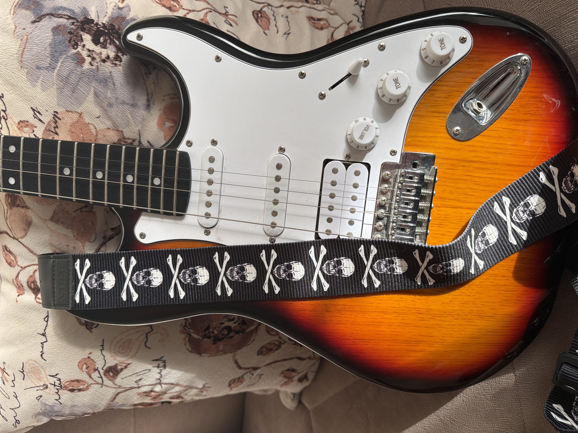 An electric guitar with a sunburst finish rests on a couch, partially covered by a pillow with a floral pattern. A black guitar strap with a skull and crossbones design is draped across the guitar. The guitar has a white pickguard, three white pickups, and chrome hardware, including volume and tone knobs. The scene is lit by natural light, casting shadows on the guitar and pillow. The location is not discernible from the image.
