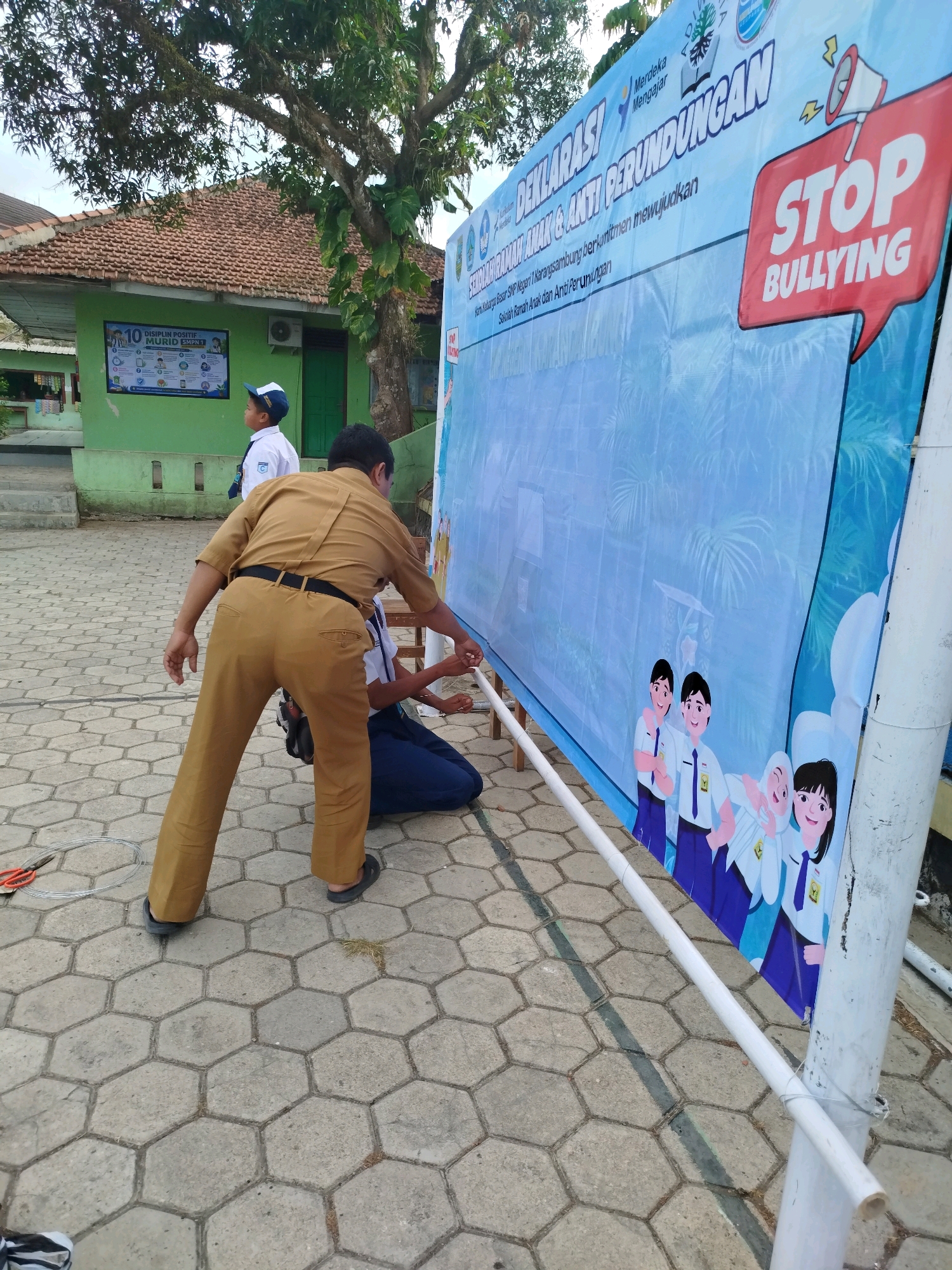 In Karangsambung, Indonesia, a group of individuals are setting up a large blue banner that reads "STOP BULLYING" in red lettering. The banner also displays cartoon depictions of students and text in Indonesian. Two people, a man in a tan uniform and a person in a blue school uniform, are actively working on securing the banner with white poles and wire. In the background, a green building with a tiled roof is visible, along with a tree and another student in a school uniform. The ground is paved with hexagonal concrete tiles.
