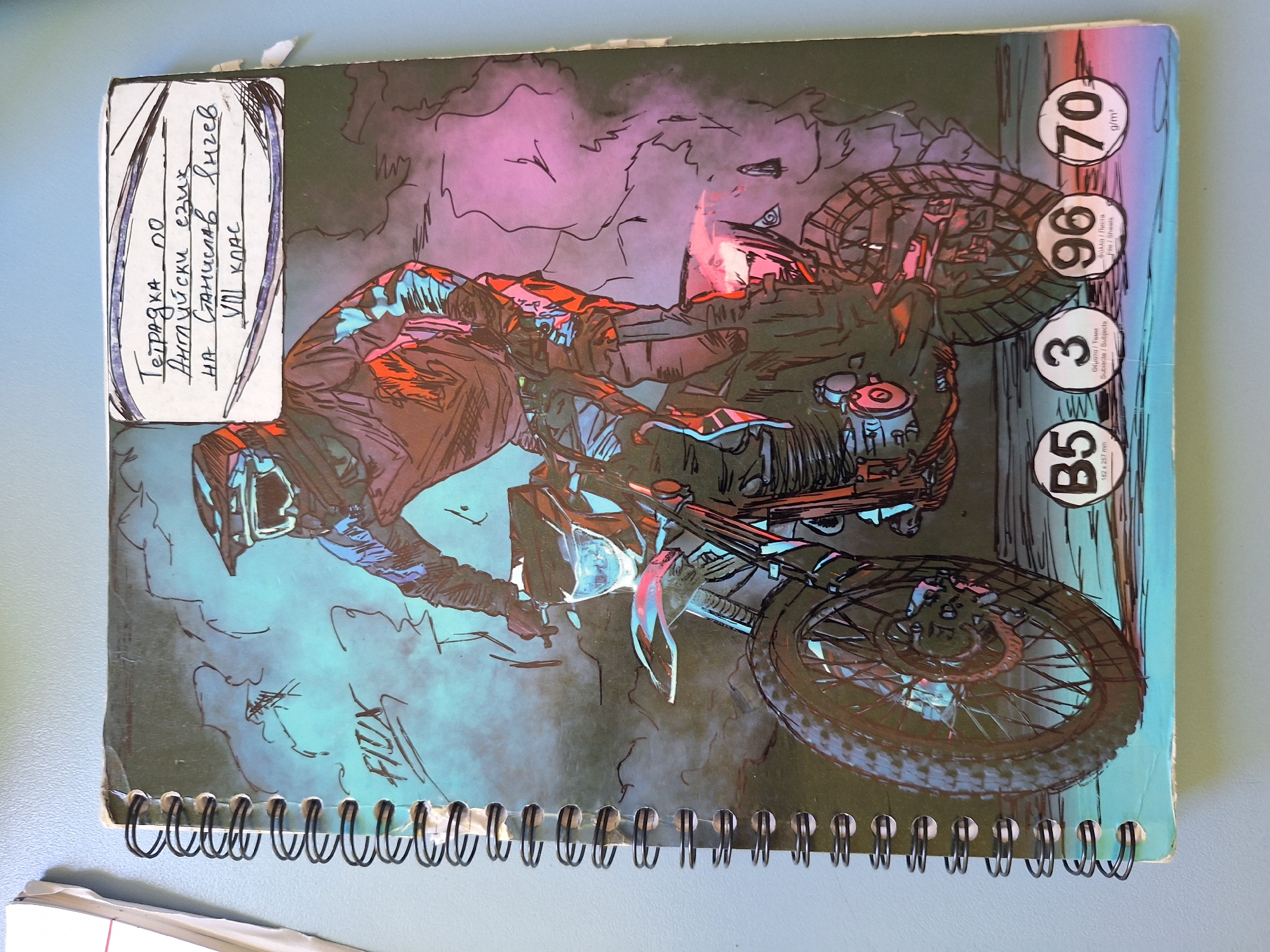 The image is a close-up, overhead shot of a spiral-bound notebook with a drawing of a motocross rider on a bike. The notebook is resting on a light blue surface, possibly a desk or table. The drawing is in a stylized, comic-book-like fashion, with bold outlines and vibrant colors, predominantly blues, purples, oranges, and blacks, creating a dramatic effect. The rider is wearing a helmet and goggles, and the motorcycle appears to be in motion, with smoke or dust effects around it.
In the upper right corner of the notebook's cover, there is a handwritten label in Bulgarian. The text reads: "Тетрадка по Английски език на Станислав Ангелов VIII клас" which translates to "Notebook for English language by Stanislav Angelov, VIII grade."
Below the drawing, there are several circular icons with text. These appear to be specifications or ratings for the paper. The icons show the numbers "B5", "3", "96", and "70". Beneath these numbers, there is text in English and what appears to be Greek, indicating paper size, number of subjects, number of sheets, and g/m² (grams per square meter). Specifically, "B5 182 x 257 mm", "3 Θέματα / Теми Subjects / Subjects", "96 Φύλλα / Листа File / Sheets", and "70 g/m²".
The overall impression is of a personalized school notebook for a student. There is no information in the image that indicates a specific location in the city of Varna, Bulgaria. The image focuses solely on the notebook and its cover art.