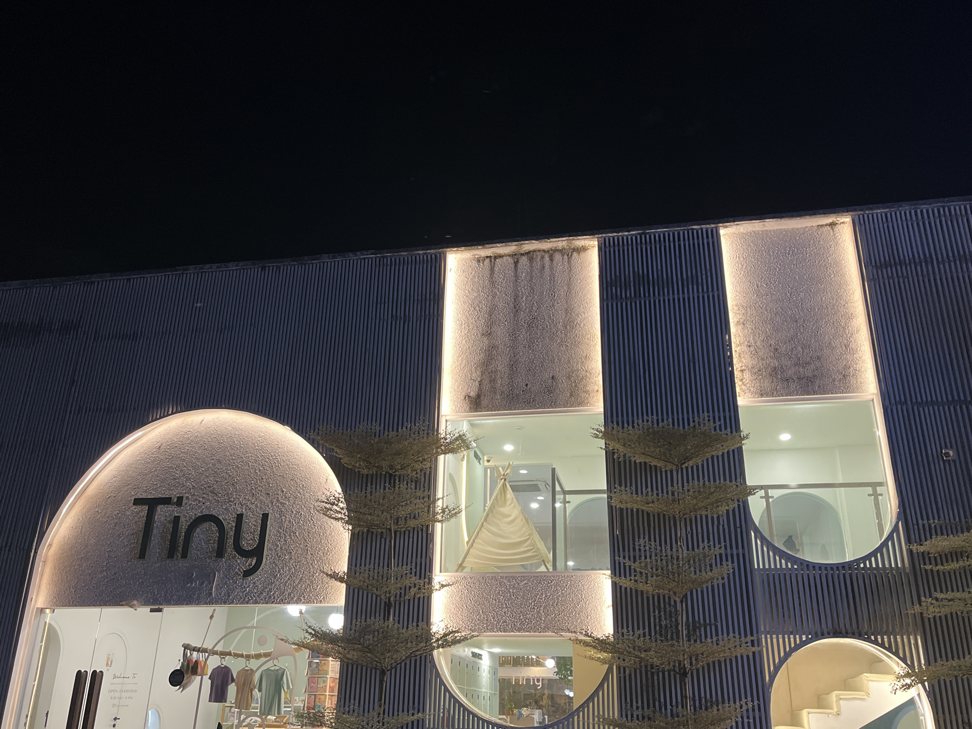 This is a night-time exterior shot of a store called "Tiny" located in North Kuta, Indonesia. The store has a modern, minimalist design with a curved, illuminated facade featuring the store's name in dark lettering. The building is characterized by vertical slats and rectangular light fixtures that highlight the textured walls. Through the large windows, a children's play area with a teepee is visible, along with clothing racks displaying colorful children's apparel. A staircase is also visible in the lower right section of the image. The overall impression is of a welcoming and stylish children's boutique.
