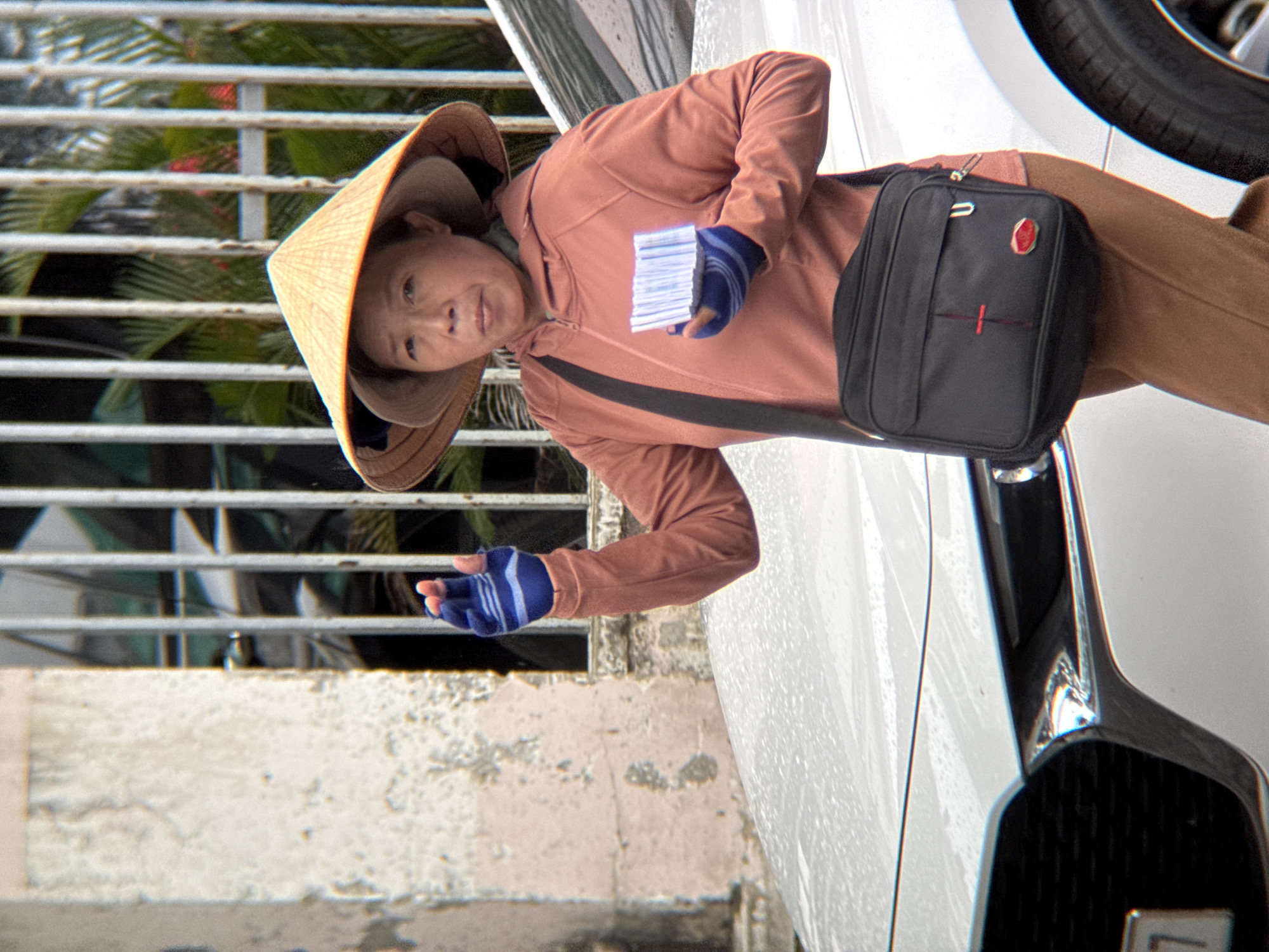 A woman wearing a conical hat and a pink jacket stands next to a white car. She has a black bag slung across her body and is holding a stack of white cards in her gloved hand. The woman's left hand is raised, palm outward, as if signaling. The background consists of a building with a barred window and foliage. The scene appears to be in a city, potentially on a street in Nha Trang, Vietnam, given the context.