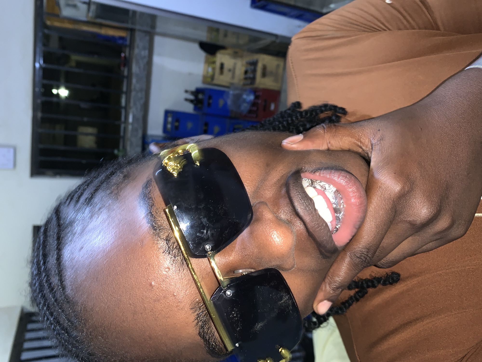 A close-up, head-and-shoulders portrait depicts an individual with dark brown skin and braided black hair, looking directly towards the viewer. The person is wearing black-lensed, gold-framed sunglasses; a gold-colored emblem, possibly a lion's head, is visible on the left temple piece. The individual is smiling broadly, revealing natural white upper teeth and lower teeth covered by a reflective, silver-colored material, consistent with a dental grill or temporary foil. The person's right hand (viewer's left) is raised, with the index finger and thumb touching the chin and lower lip area. The individual is wearing a brown, short-sleeved or long-sleeved shirt with visible ribbed texture, and a metallic, possibly silver-colored, bracelet on their right wrist (viewer's left).
The background is out of focus, indicating an indoor setting. In the upper background, a window with black vertical and horizontal security bars is visible, with a single bright, circular light source visible through the bars. To the right, stacks of various colored plastic crates (blue, red, and yellow/brown) are present, some appearing to contain bottles. Walls are light-colored. No discernible signage or specific architectural features are present within the image itself to confirm the location as New Stadium, Nigeria.