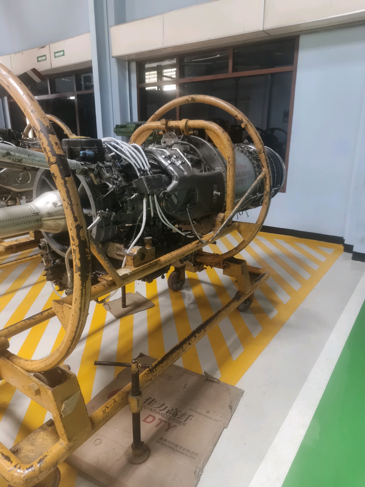 The image shows an aircraft engine on a yellow metal stand with wheels. The engine is complex, with many tubes and wires connected to it. The stand is designed to hold the engine securely and allow it to be moved. The engine appears to be in a workshop or hangar, with concrete floors and a white wall in the background. There are yellow and white striped markings on the floor, indicating a designated area. The overall impression is one of a functional, industrial environment where aircraft maintenance or repair is carried out. The image is taken from a slightly low angle, looking up at the engine. There are no people visible in the image.