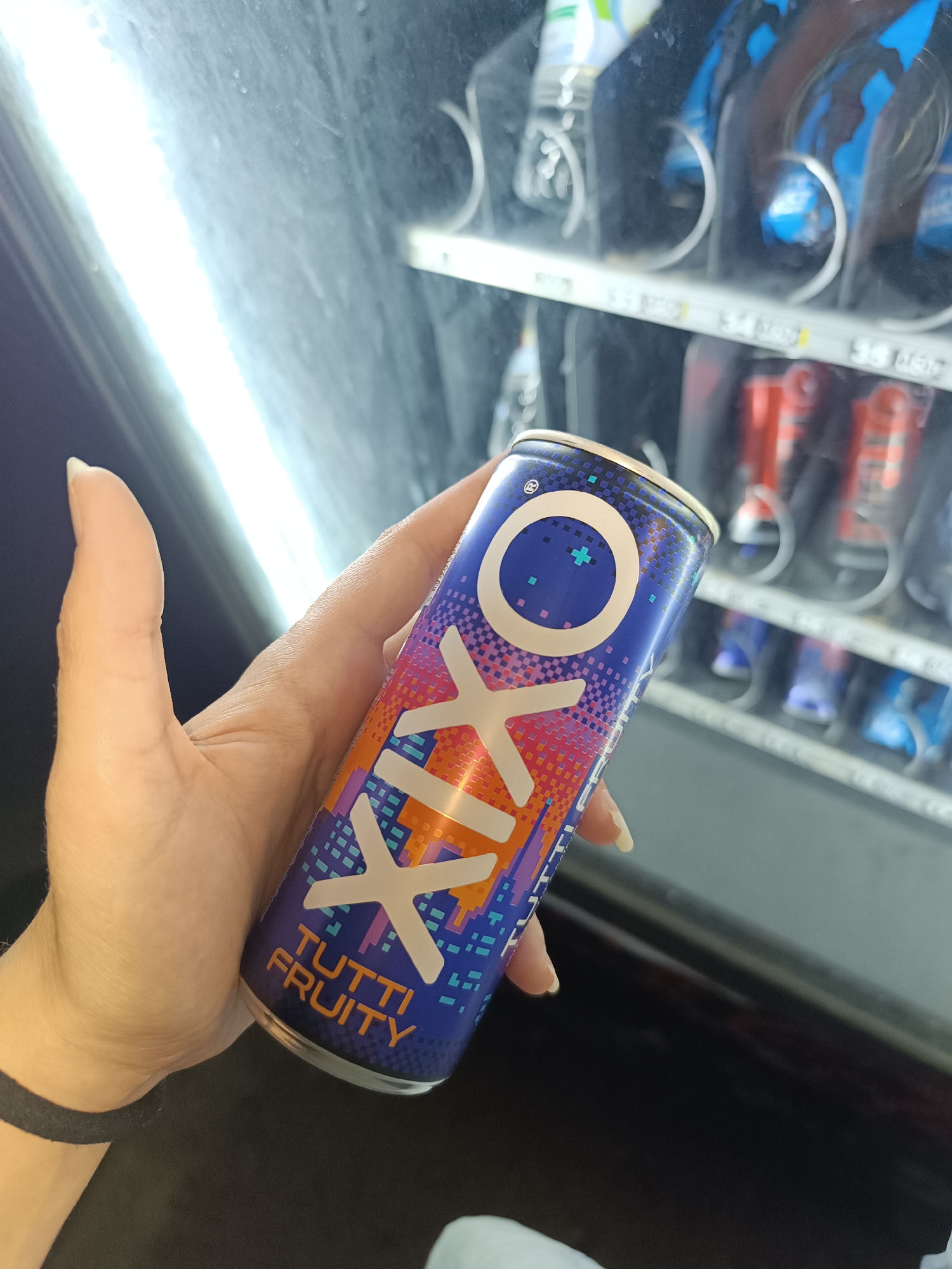 A hand holds a can of "OXXO Tutti Fruity" energy drink in front of a vending machine. The can is primarily blue with orange and white accents, displaying the "OXXO" logo. The vending machine is stocked with various drinks. The location is inferred to be Pleven, Bulgaria, based on the presence of the "OXXO" brand, which has a market presence in Bulgaria.