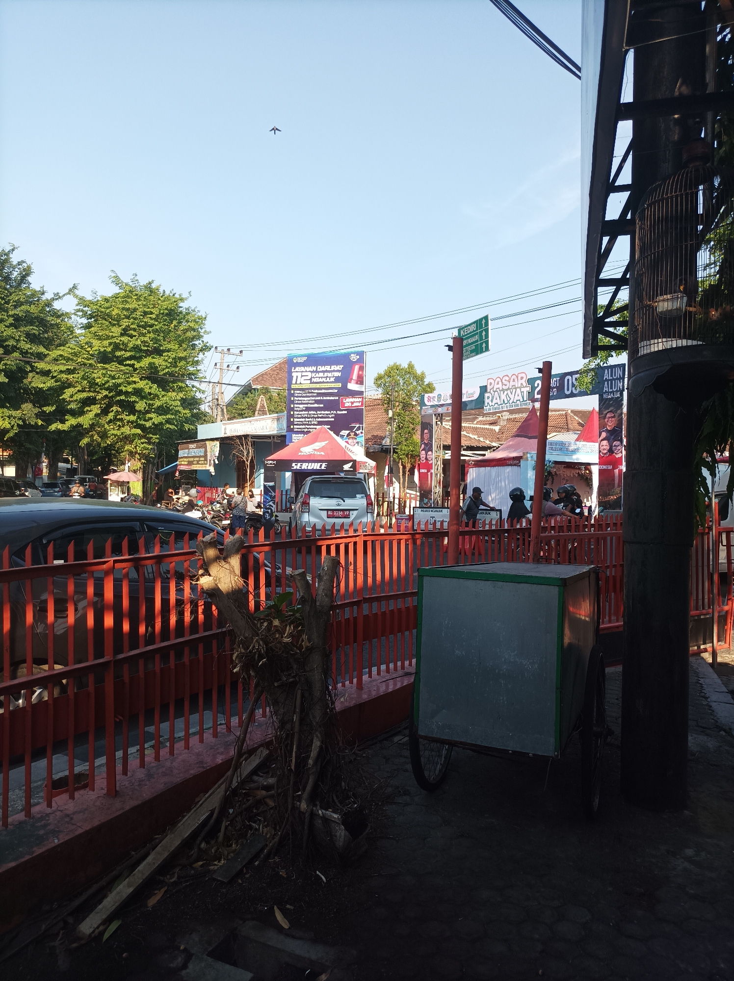 The image depicts a street scene in Nganjuk, Indonesia, on a clear day under a bright blue sky. In the foreground, a metal cart with green trim is parked on a paved surface. To the left, an orange metal fence runs horizontally, behind which are several parked cars and lush green trees. Further down the street, there are tents set up, suggesting an outdoor market or event. Several people are visible, some on motorcycles and others standing. Prominent banners and signs are displayed, including one that reads "PASAR RAKYAT 2" (People's Market 2) and others with text and images of people, likely advertising local government services or political campaigns. A traffic sign pointing to "KEDIRI" and "ALUN ALUN" is also visible. In the background, buildings and utility poles are present. A small bird is captured in flight against the sky. To the far right, a tall, dark cylindrical structure is visible, with a birdcage hanging from it, containing a bird.
