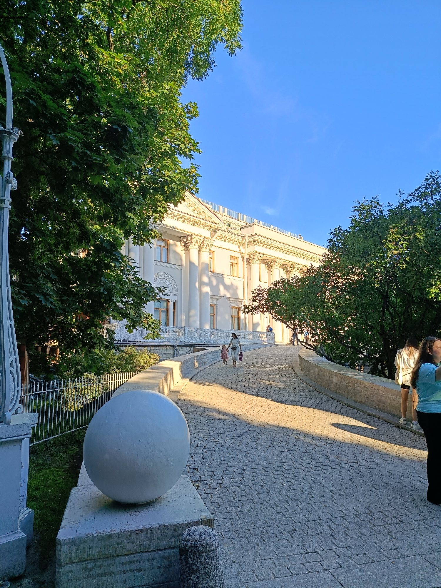 The image depicts a paved pathway leading towards a neoclassical building with a grand facade, featuring columns and an ornate roofline. Lush green trees with dense foliage frame the scene on both sides, with sunlight filtering through the leaves, casting dappled shadows on the pathway. In the foreground, a large, smooth white sphere rests on a stone pedestal. Several people are visible on the pathway, including a woman holding a child's hand as they walk away from the camera, and a few individuals standing further up the path near the building. The sky is a clear, bright blue, suggesting a sunny day. The pathway is constructed of grey cobblestones, and a low stone wall borders its edge. A white decorative fence is present on the left side of the pathway, along with a portion of a white lamppost.