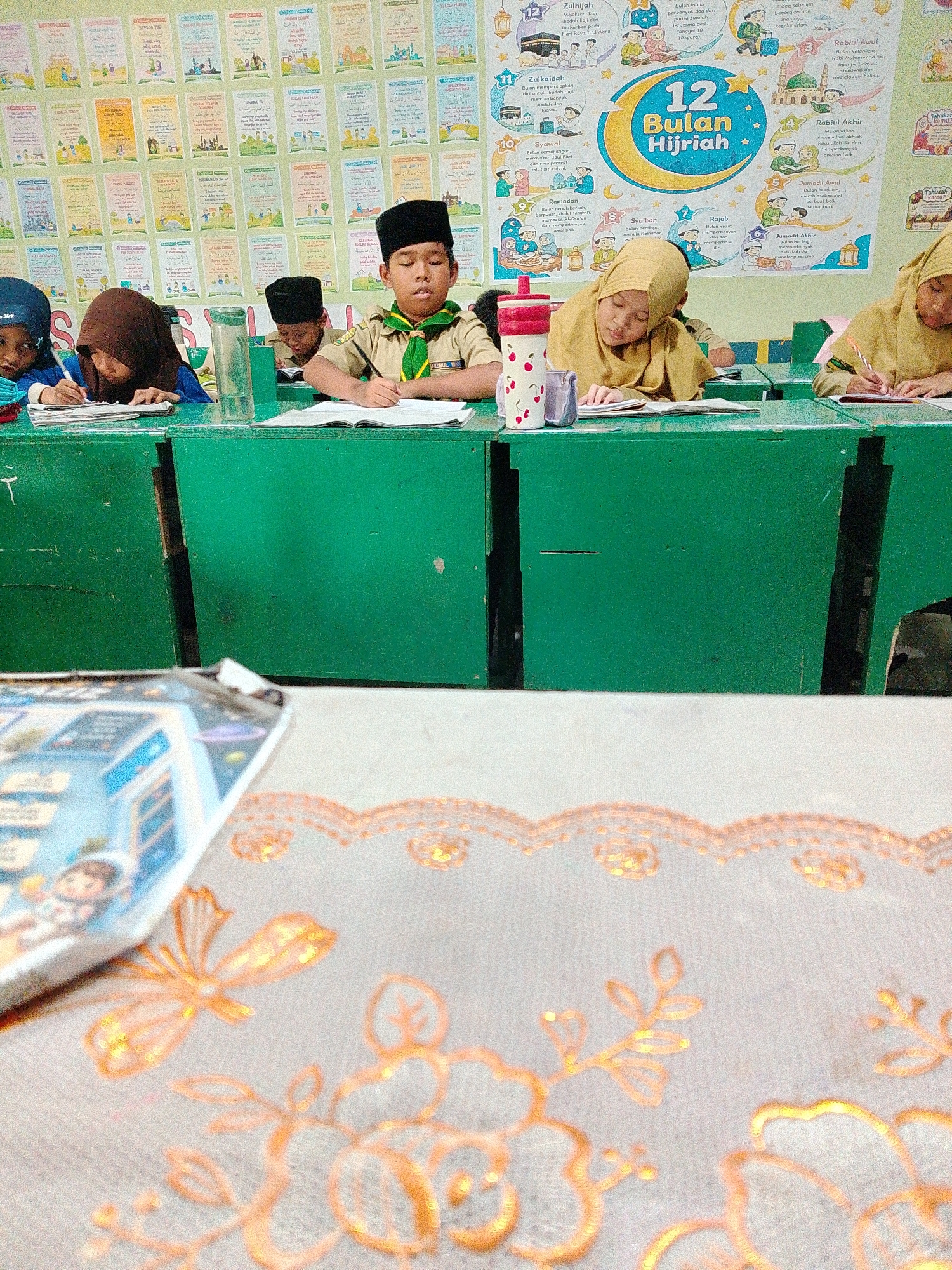 This is a medium shot, eye-level photograph taken indoors, likely a classroom in Tembung, Indonesia. The foreground is dominated by a blurred white fabric with an ornate, orange embroidered pattern of flowers and leaves, and a partially blurred magazine or book with a picture of a cartoon astronaut on it. The middle ground features a long, bright green wooden table or desk. Seated behind the desk are four children, three girls and one boy, all appearing to be of elementary school age.
On the left, a girl wearing a dark blue hijab is focused on writing in a notebook. Next to her, another girl in a brown hijab is also writing. In the center, a boy wearing a brown and green scout uniform with a black peci hat is actively writing in an open book, holding a pen in his right hand. To his right, a girl in a light yellow hijab is also engaged in writing. Several objects are visible on the desk, including a red and white patterned water bottle with a straw, a glass of water, and various notebooks and papers.
The background consists of a wall covered with educational posters. These posters display Islamic themes, including calligraphy, drawings of mosques, and information about the "12 Bulan Hijriah" (12 Hijriah Months), illustrated with moons, stars, and scenes depicting Islamic traditions like Hajj and Ramadan. The posters are colorful and appear to be in Indonesian and Arabic. The overall scene depicts a classroom setting where students are engaged in learning activities.