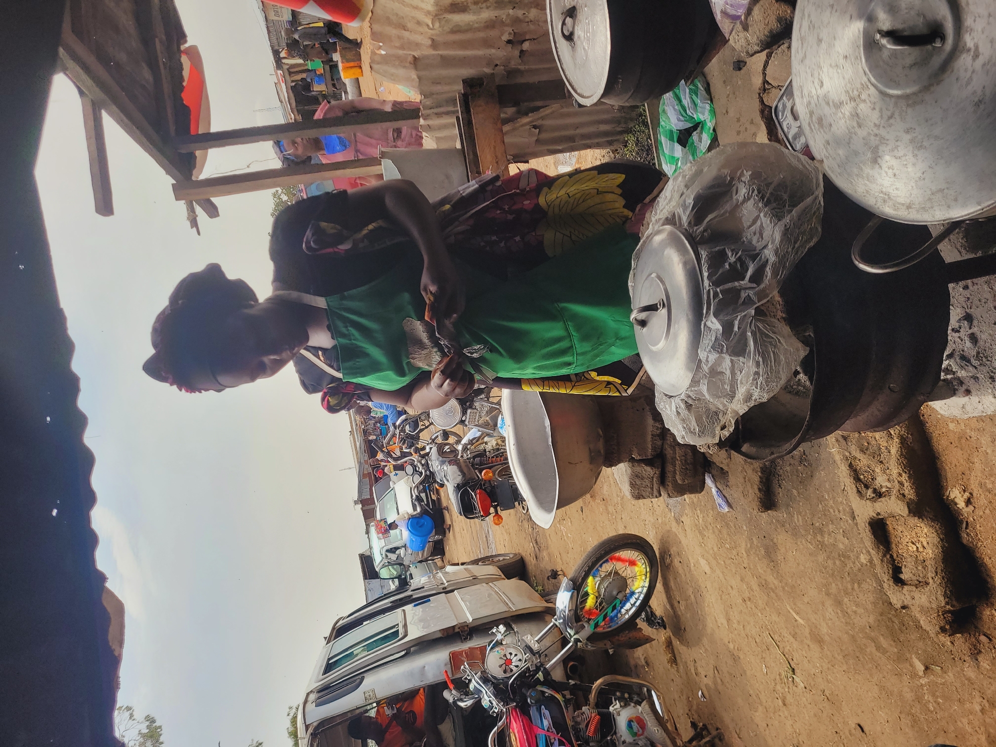 A woman wearing a green apron over a patterned dress and headwrap stands in a market environment in Bukuru, Nigeria. She is looking down at something she is holding in her hands. In front of her are several large cooking pots, some covered with plastic wrap. Behind her, a silver van is parked, with its door open and a person sitting inside. Various motorcycles, a large metal basin, and other market stalls and structures are visible in the background, indicating a busy outdoor marketplace. The ground is dirt and sand. The sky is overcast.