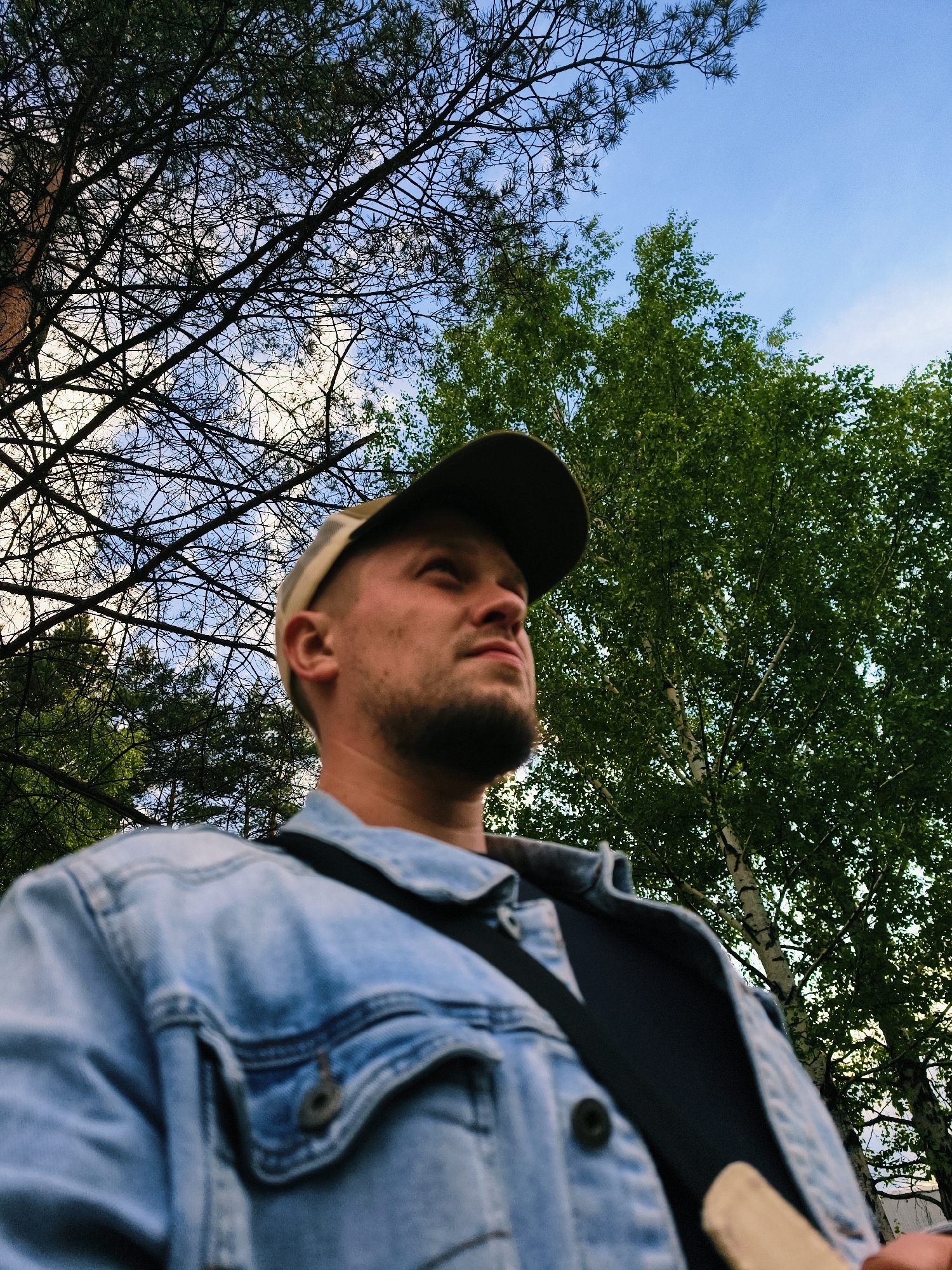 The image is a low-angle shot of a man with a beard wearing a denim jacket and a baseball cap. A strap is across his torso. He appears to be looking up. Behind him are trees and a sky with some clouds. The location is likely Noviy, Russia.