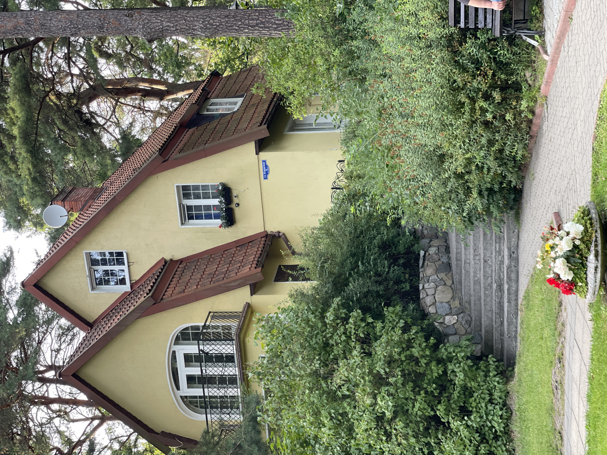 The image depicts a yellow house with a red tiled roof, nestled among trees in Svetlogorsk, Russia. The house features an arched balcony with a wrought-iron railing on the second floor, and several windows with white frames. A satellite dish is mounted on the roof, and a street sign reading "ул. Курортная" (Resort Street) is visible on the house's exterior. In the foreground, a stone staircase leads up to the house, bordered by lush green bushes. A paved walkway runs alongside the bushes, and a park bench is visible on the right side of the frame. A planter with red and white flowers sits on the grass in front of the walkway. The scene is bathed in natural light, suggesting it was taken during the daytime.