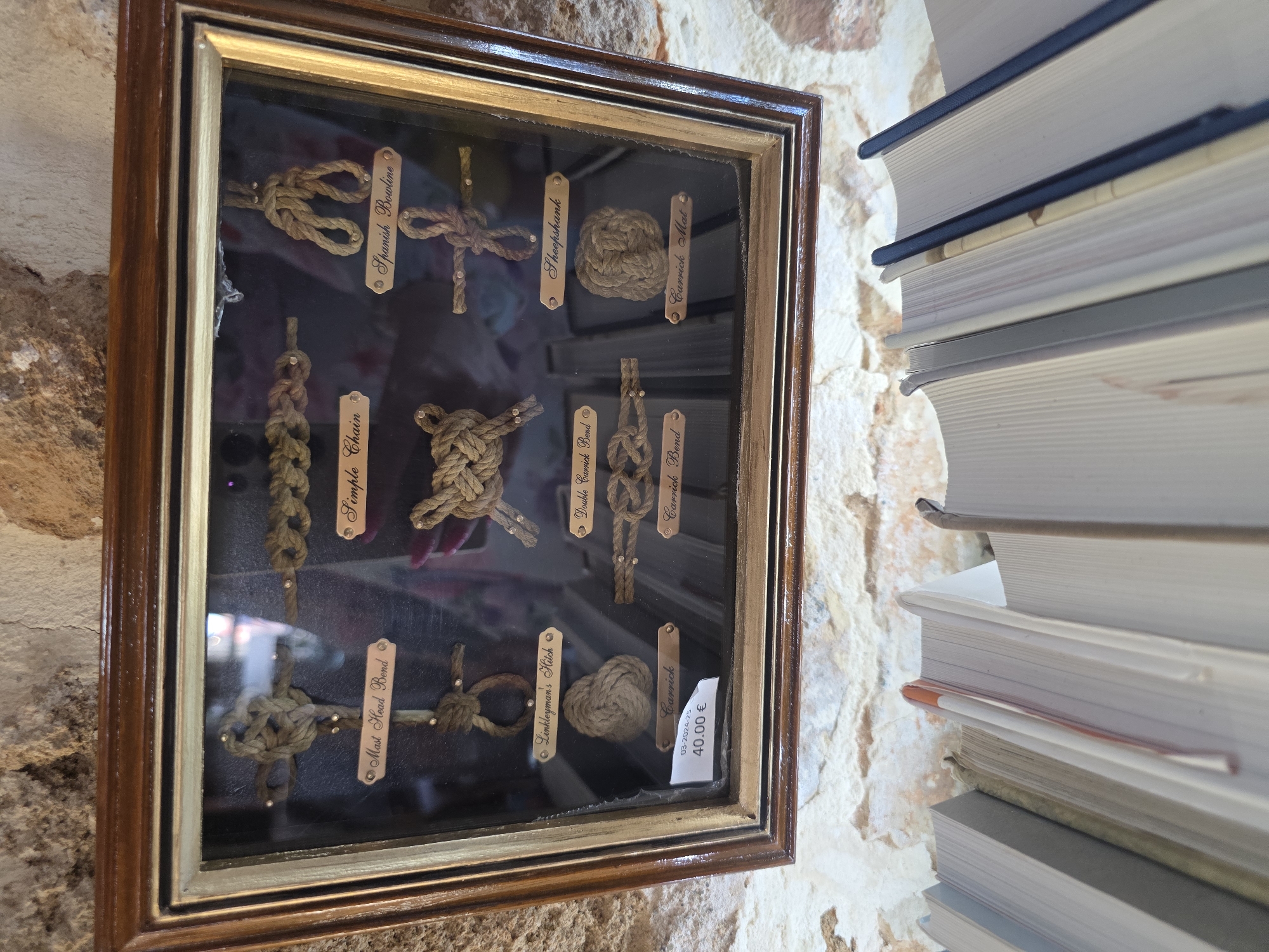 The image shows a framed display of sailor's knots, likely a souvenir or educational item. The frame is made of dark wood with a gold inset. Inside the frame, there are nine different knots made of rope, each with a small plaque identifying the knot by name. The knots are arranged in three rows of three. Some of the identified knots are "Mast Head Bend," "Simple Chain," "Spanish Bowline," "Linkleyman's Hitch," "Sheepshank," "Carrick Bend," and "Carrick Mat." A price tag is visible in the bottom left corner, showing "40.00 €" and a date "03-2024-25". The display is set against a textured stone wall, and in the foreground, a row of books is visible. Based on external information, this display is located in Chania, Greece.