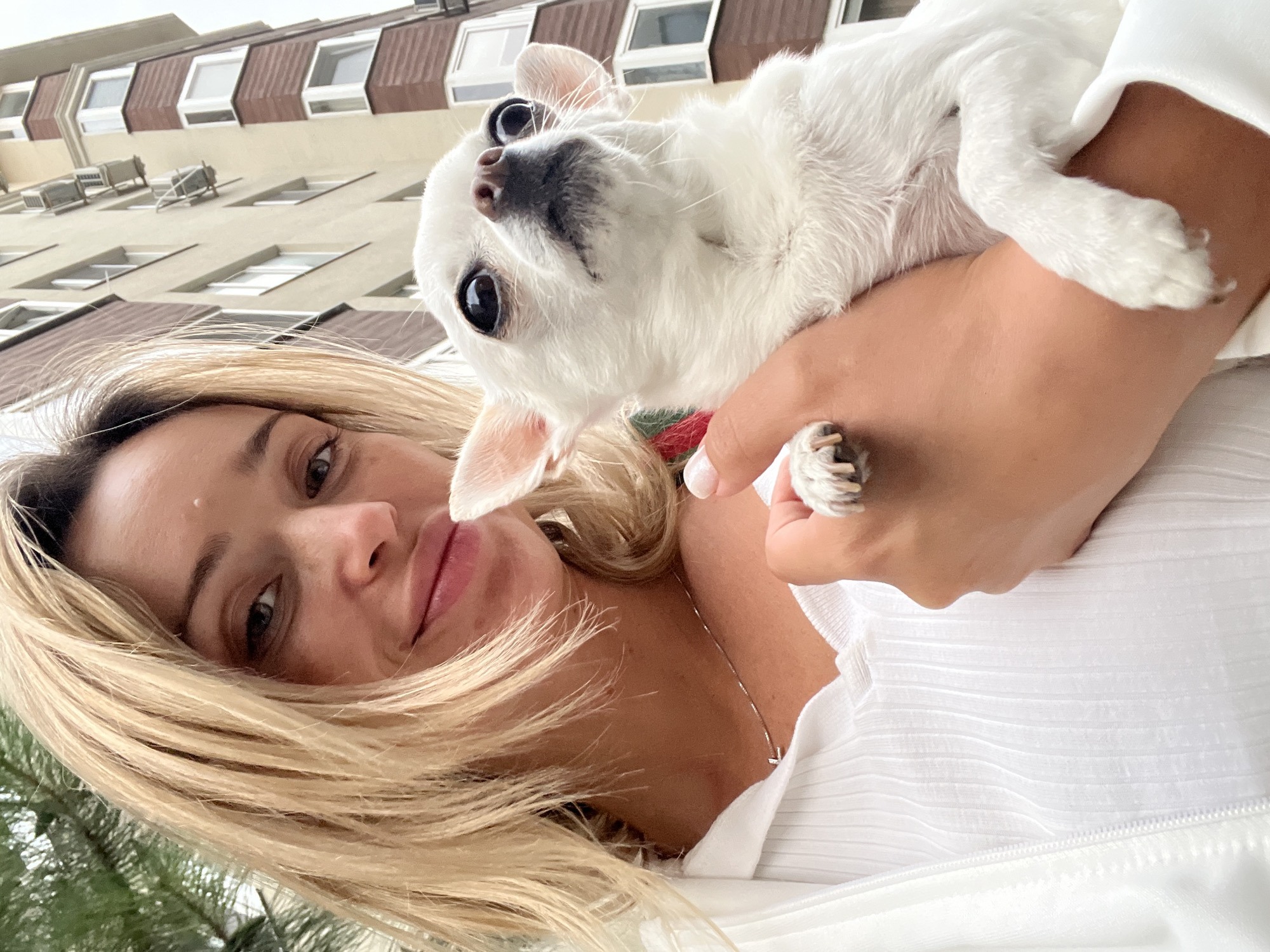 A blonde woman with fair skin and light freckles is taking a selfie outdoors, holding a small white chihuahua. The woman is wearing a white ribbed top and a white open jacket. A thin silver necklace is visible around her neck. The chihuahua has large dark eyes and big ears, and it is looking upwards and slightly to the right. The background shows a multi-story building with a light beige facade and brown trim. There are several window units for air conditioning attached to the building. Trees with green foliage are visible on the left side of the image, and part of a building's exterior is seen on the right. The scene appears to be set in an urban environment, possibly a residential area. The location is identified as Kryzhanivka, Ukraine.