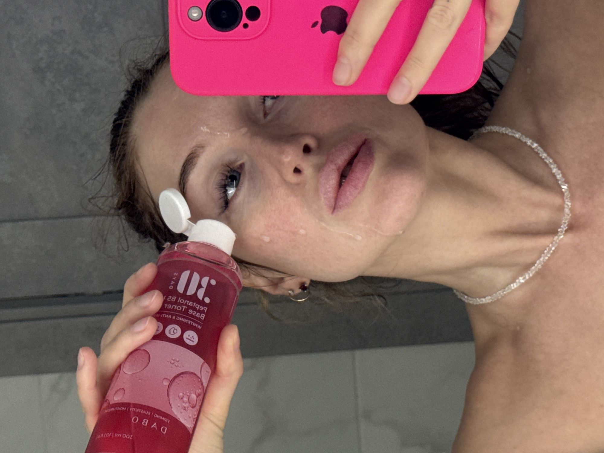 A woman is in a bathroom, likely taking a selfie with a pink iPhone. She is holding a pink bottle of "DABO 30 Peptotalk Base Toner" in her left hand and is wet, presumably from washing her face. The background appears to be a gray tiled wall. The location in the city of Chaikovsky, Russia, is not discernible from the image.
