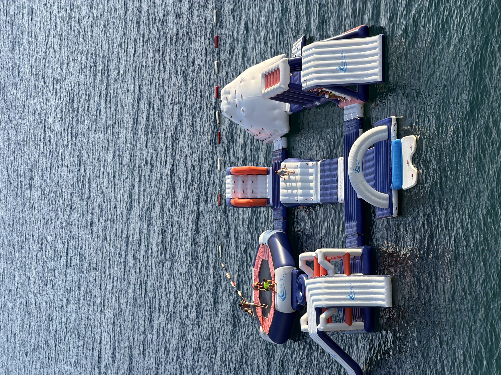 An aerial, high-angle shot captures a floating inflatable water park situated in the calm, deep blue waters of the sea, likely near the city of Boreti, Montenegro. The water park is a vibrant collection of interconnected inflatable structures, primarily white and blue with orange accents. These structures include a large trampoline-like platform with at least three individuals on it, one actively jumping. Adjacent to this are various climbing walls, slides, and obstacle courses, all designed for water recreation. Several people are visible on different parts of the inflatable park, engaged in activities. In the background, a line of red and white buoys demarcates a boundary in the water. The overall scene is one of summer leisure and aquatic fun.