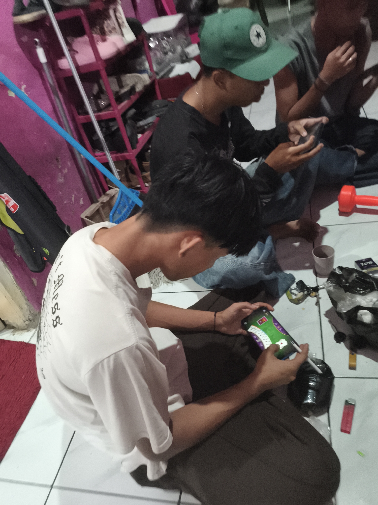 The image depicts a group of young males in a room with tiled flooring, possibly an indoor recreational area or living space, located in Depok, Indonesia. The lighting is dim, suggesting it may be evening or nighttime. One individual in the foreground is wearing a white t-shirt with a graphic design on the back and dark pants. He is seated on the floor, engrossed in playing a mobile game on his smartphone. Another individual, wearing a black long-sleeved shirt and a green baseball cap, is also seated and appears to be playing a game on his phone. A third person, partially visible, is also present. Scattered on the floor are various objects including a red plastic object resembling a plunger or toy, a white paper cup, a small black plastic bag, a cigarette pack, and a red lighter. A blue rope or cord is visible in the background, along with a pink shelving unit and a black bag. The overall scene suggests a casual gathering where the individuals are engaged in personal activities, primarily using their mobile phones.