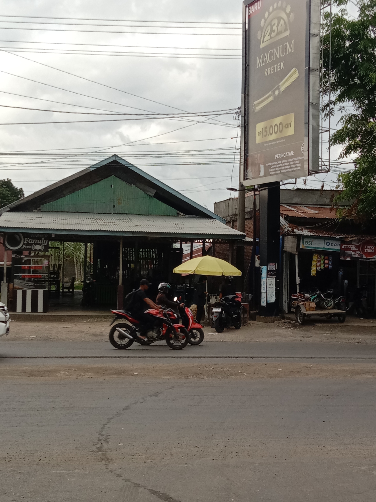 The image shows a street scene in what appears to be Lhokseumawe, Indonesia. Two individuals on motorcycles are the focal point, with the motorcycle closer to the camera being red and black.
Several businesses line the street. To the left is a structure with a green roof, labeled "Warkop Family Free" - indicating a local coffee shop or gathering place. Further down the street, there's a commercial establishment advertising BSI, ODAB, and Lucky Strike cigarettes. Next to this is a yellow tent and more motorcycles.
Dominating the right side of the frame is a large billboard advertising "234 Magnum Kretek" cigarettes, with a price of "RP15,000-an." The sky is overcast, and power lines are visible overhead. The street is paved but appears to be worn.