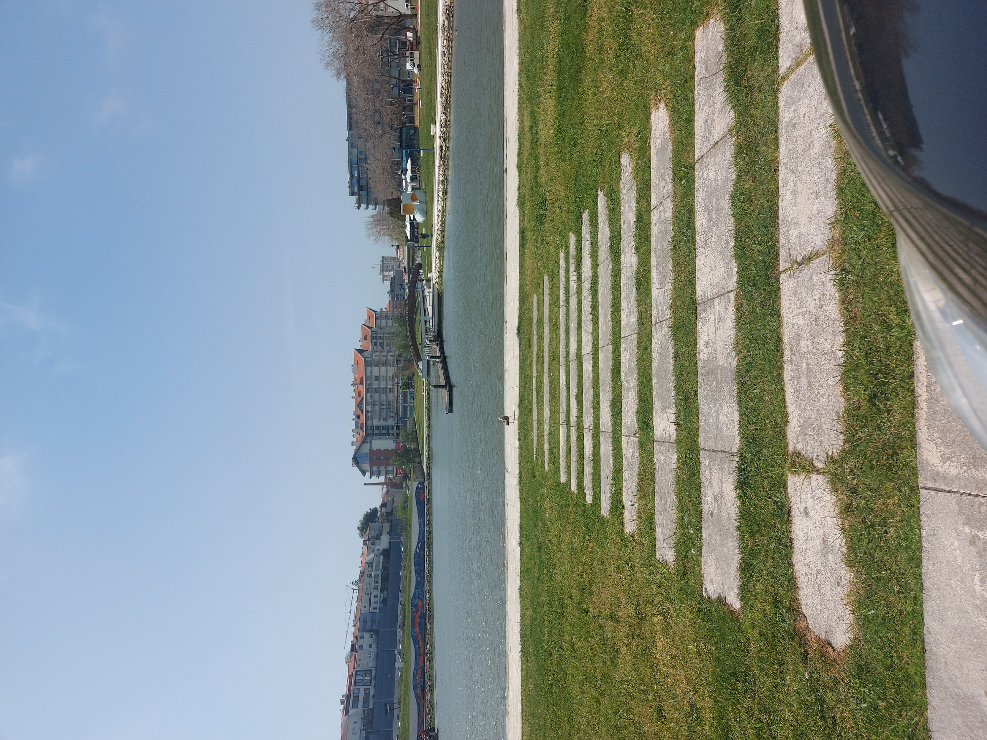 The image shows a waterfront area in Aveiro, Portugal, featuring a walkway with alternating concrete slabs and grassy areas. The waterway is visible to the left of the walkway, with buildings lining the opposite bank. The sky is clear and blue. A seagull is standing by the water's edge. A structure of blue and orange tiles runs parallel to the waterfront along the waterway.