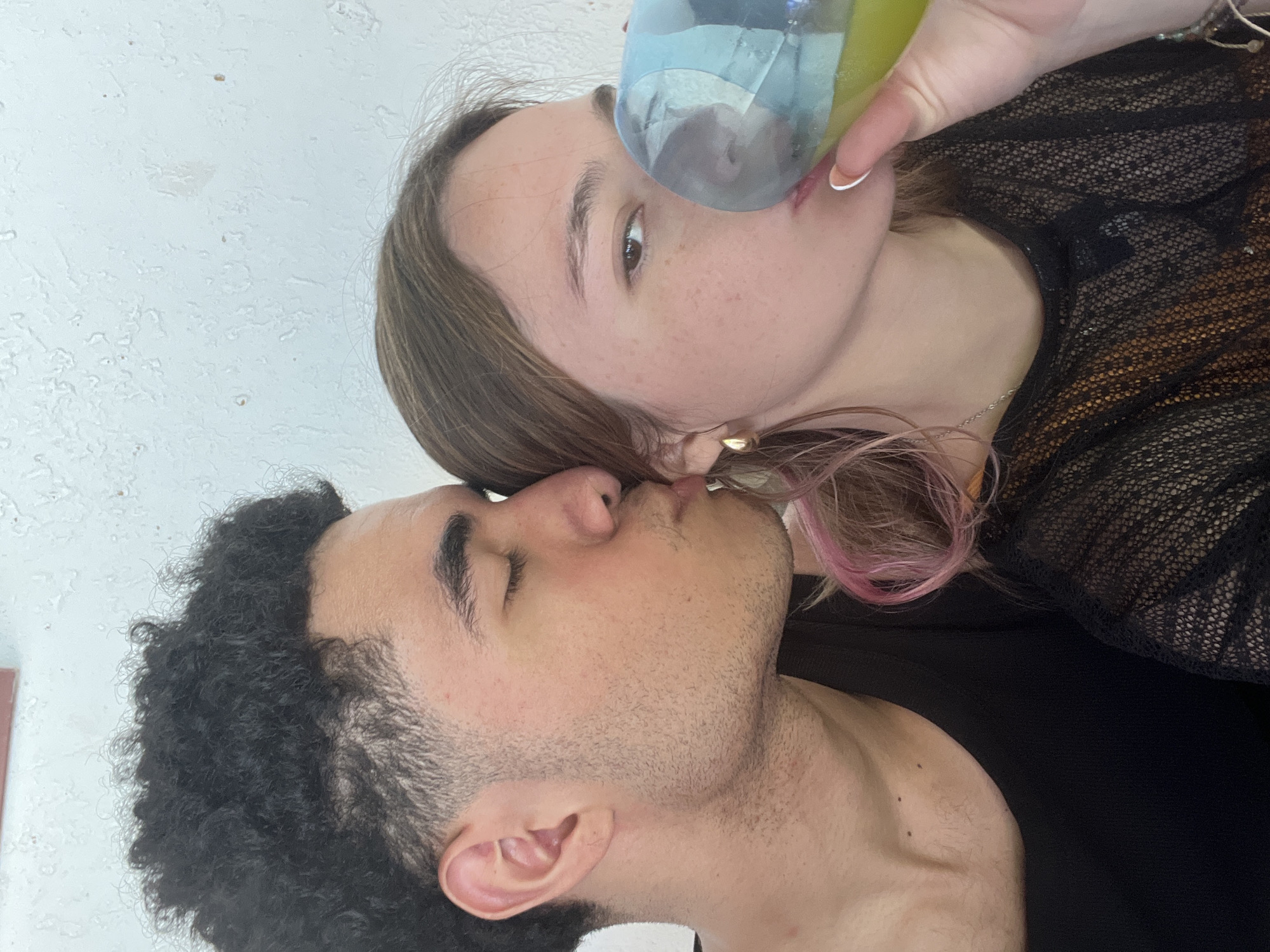 The image depicts two individuals in an indoor setting. The woman is holding a water bottle to her mouth and has brown hair with pink highlights, wearing a black lace top. The man has short, curly black hair and stubble, wearing a black shirt. They appear to be kissing. The location is Unknown, Unknown.