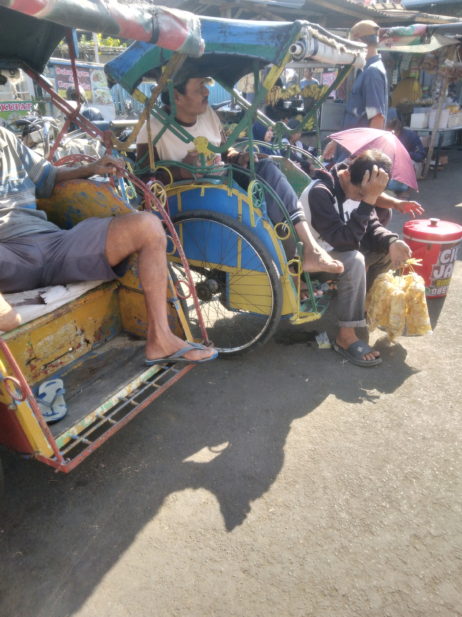 The image shows a street scene in Sokaraja, Indonesia, with a brightly colored becak (pedicab) as the central focus. Three men are visible. The driver of the becak is seated and looking to his right. A second man is seated behind the driver, appearing to be a passenger, and is holding his head with his left hand. He is also holding a bunch of yellow food items in his right hand. A third man, wearing a blue shirt, stands to the right of the becak, looking to his left. The becak itself is yellow and blue with red trim, and it has a large wheel visible. In the background, there are stalls with awnings, suggesting a market or roadside vendors. The ground is asphalt, and shadows are cast by the becak and people, indicating a sunny day. A red cooler is also visible on the ground.