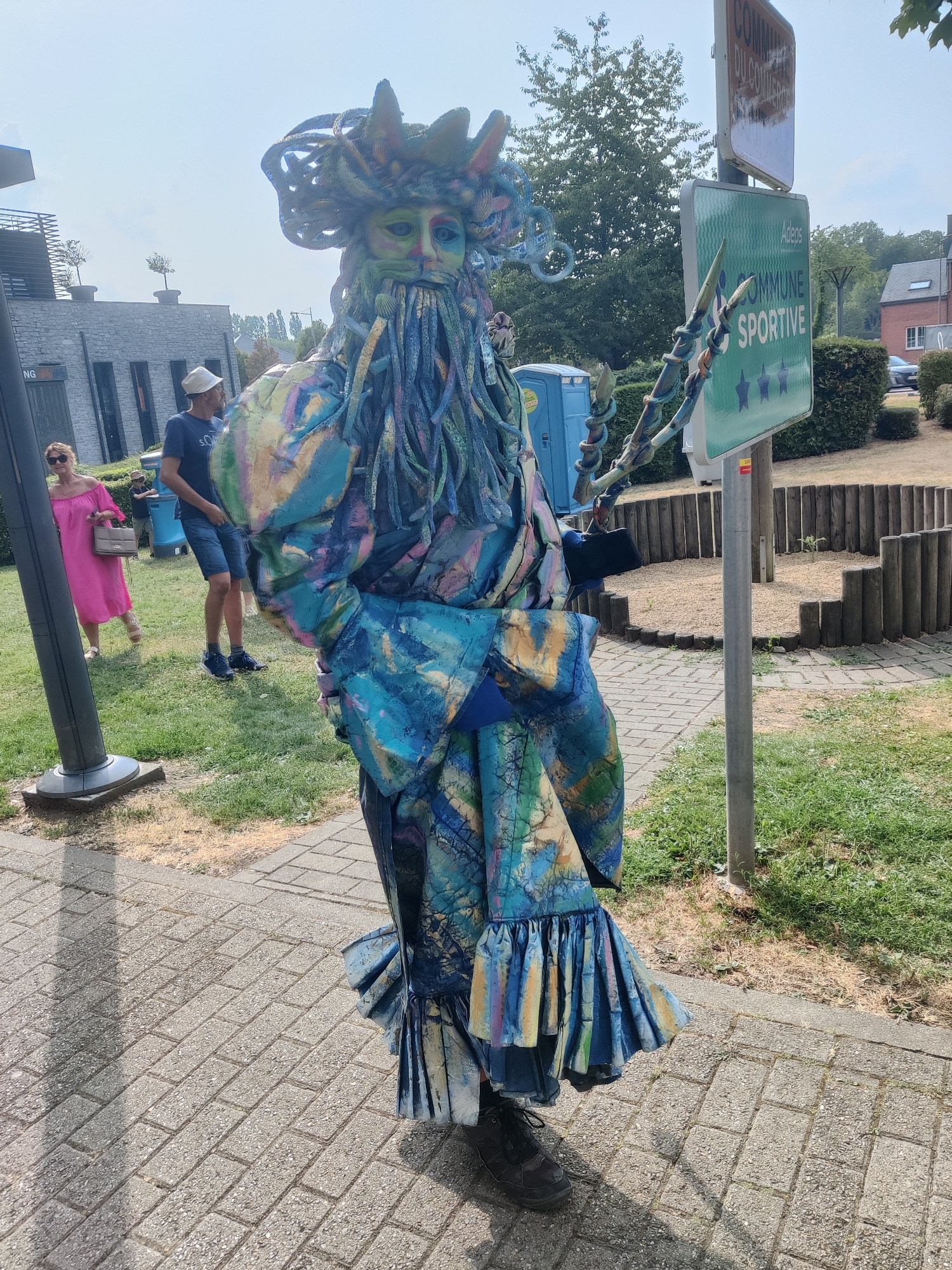 A costumed individual, dressed in a vibrant, multi-colored outfit with an elaborate headpiece and long, flowing beard, stands on a paved walkway. The costume features a patterned fabric in blues, greens, yellows, and purples, with a ruffled skirt. The individual is holding a staff-like object in their right hand. In the background, a woman in a bright pink dress walks on a grassy area. A man wearing a white hat and a blue t-shirt stands near her. A blue portable toilet is visible further in the background, along with buildings and trees. A green sign with white text and stars is mounted on a pole. The scene appears to be outdoors on a sunny day, likely in a public space within the commune of Marche-en-Famenne, Belgium.