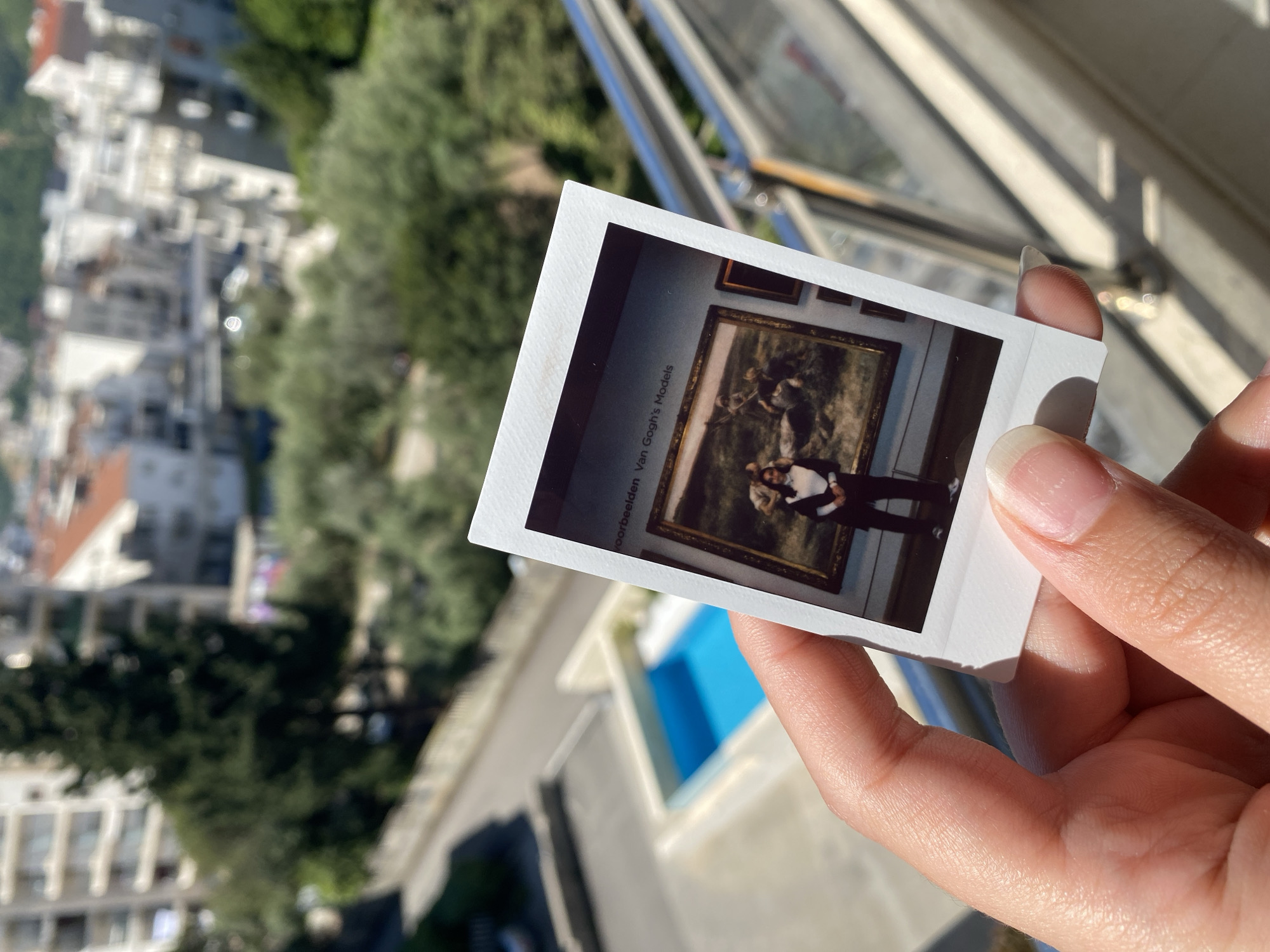 A close-up, angled shot features a hand holding a white-bordered instant film photograph, set against a blurred outdoor background of Budva, Montenegro. The hand, with natural nails, gently grips the photo, which is positioned in the lower-right part of the frame. Bright daylight illuminates the scene, casting distinct diagonal shadows from a window frame or railing onto the surface behind the hand, indicating a sunny day.
The out-of-focus background reveals a picturesque Mediterranean cityscape with white buildings, some with reddish roofs, interspersed with dense green trees. A distinct rectangular patch of vibrant blue suggests a swimming pool below, viewed from a high vantage point like a balcony.
The instant photograph itself displays an interior museum scene. A person, dressed in dark trousers and a white shirt layered with a black sleeveless top or vest, stands posing in front of a large, classical painting framed in gold. Other smaller framed artworks are partially visible on the museum wall. Vertically oriented text on the left edge of the instant photo reads: "voorbeelden Van Gogh's Models," indicating the painting or exhibition's theme. The overall image captures a cherished travel memory being revisited outdoors.