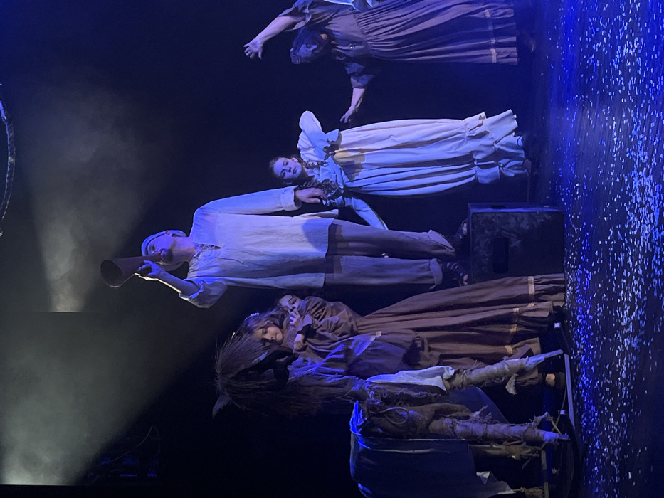 This image captures a theatrical performance on a stage in Novoshakhtinsk, Russia. Five individuals are present under blue-toned stage lighting, with a dark background and the stage floor covered in small white fragments resembling confetti or artificial snow.
Centrally, an adult male stands elevated on a dark, rectangular prop box, wearing a light-colored tunic, light trousers, and a cap. He holds a brown conical horn to his mouth, tilted upwards, as if making an announcement. To his immediate left stands a young female, approximately 6-10 years old, in a long, dark-brown dress with light trim, her hands clasped. Beside her, on the far left, is a rustic, light-colored donkey-shaped prop with a shaggy mane, mounted on a rocker base.
To the right of the central male, a young female, approximately 12-18 years old, stands in a light-cream long-sleeved dress with ruffled details, her right arm slightly raised and bent. Her gaze is directed forward. Further to the right and slightly behind, an adult female in a dark-brown long-sleeved dress performs a dramatic gesture with her left arm raised high and bent. A fifth individual, also in a dark-brown dress, is partially visible behind the gesturing female on the far right. The overhead lighting creates atmospheric haze and distinct shadows.