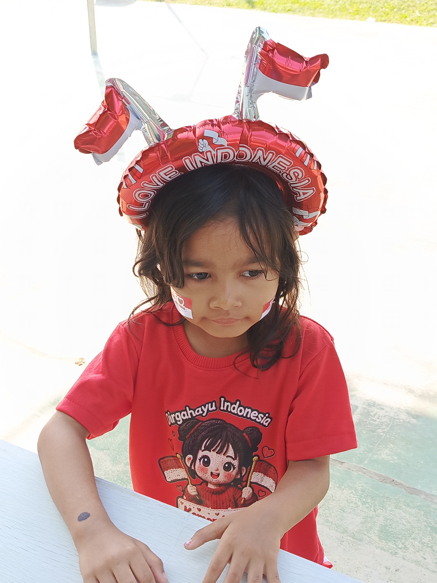 In Ciseeng, Indonesia, a young girl, likely of Indonesian ethnicity given the nationalistic imagery, is positioned outdoors, leaning on a light-colored table. She wears a red t-shirt with a graphic of a cartoon character holding an Indonesian flag and the text "Dirgahayu Indonesia" (Happy Anniversary Indonesia). Her face is adorned with small Indonesian flags on her cheeks, and she wears a festive red and silver headband with balloons shaped like Indonesian flags and the words "LOVE INDONESIA." The background consists of an outdoor paved area with some greenery visible in the distance, suggesting a park or public space. The lighting indicates it is daytime, likely sunny. Her hands are resting on the table, and her gaze is directed downwards, perhaps in thought or shyness.
