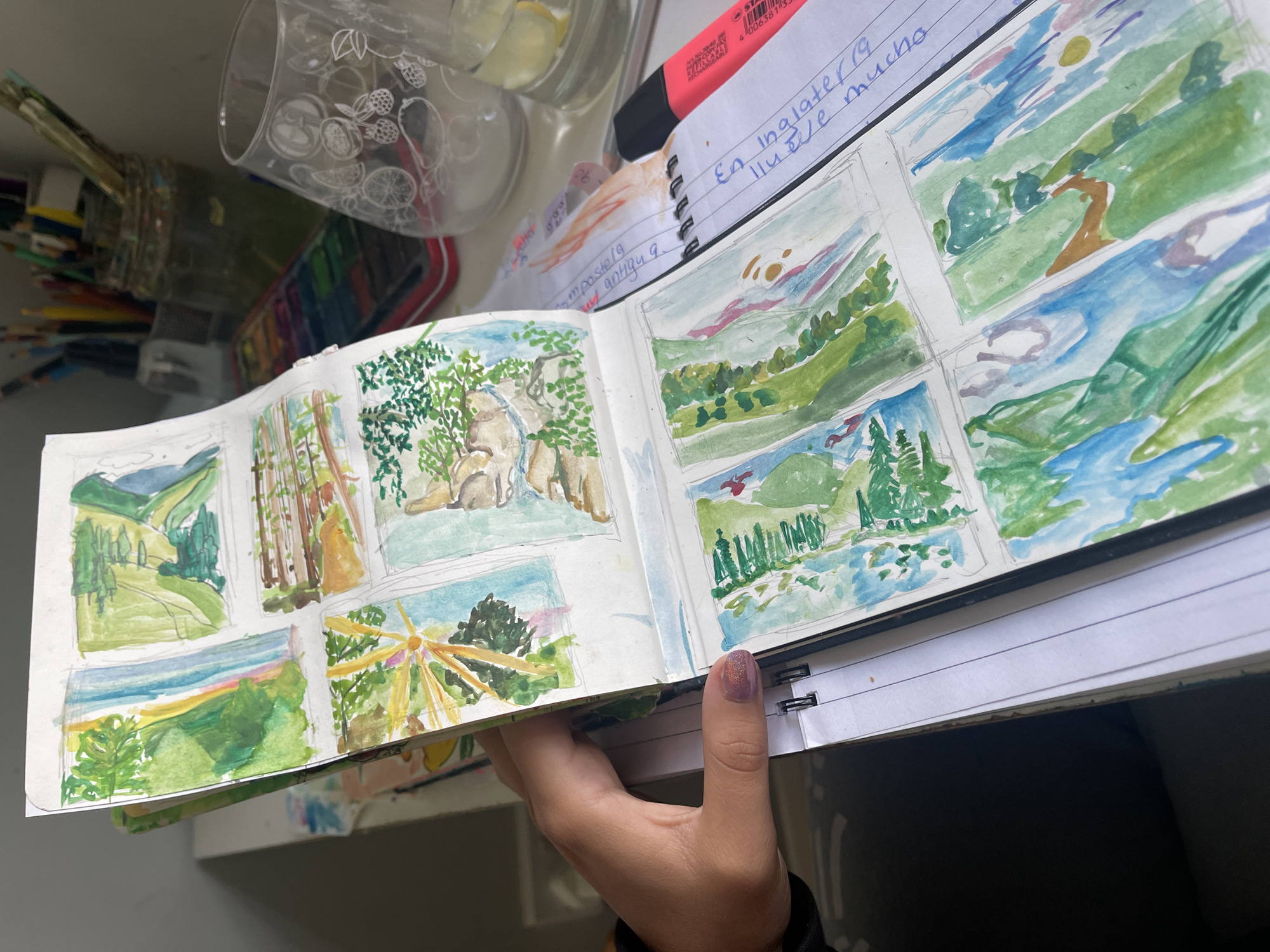 A hand is holding open a sketchbook filled with watercolor landscape paintings. The paintings depict various scenes including trees, hills, bodies of water, and architectural structures. The sketchbook is resting on top of notebooks and a desk. Miscellaneous art supplies, including a jar of paintbrushes, a watercolor palette, and colored pencils, are scattered around the sketchbook. The setting appears to be an indoor workspace, likely a desk or table. The location in Maidstone, United Kingdom is not discernible from the image.