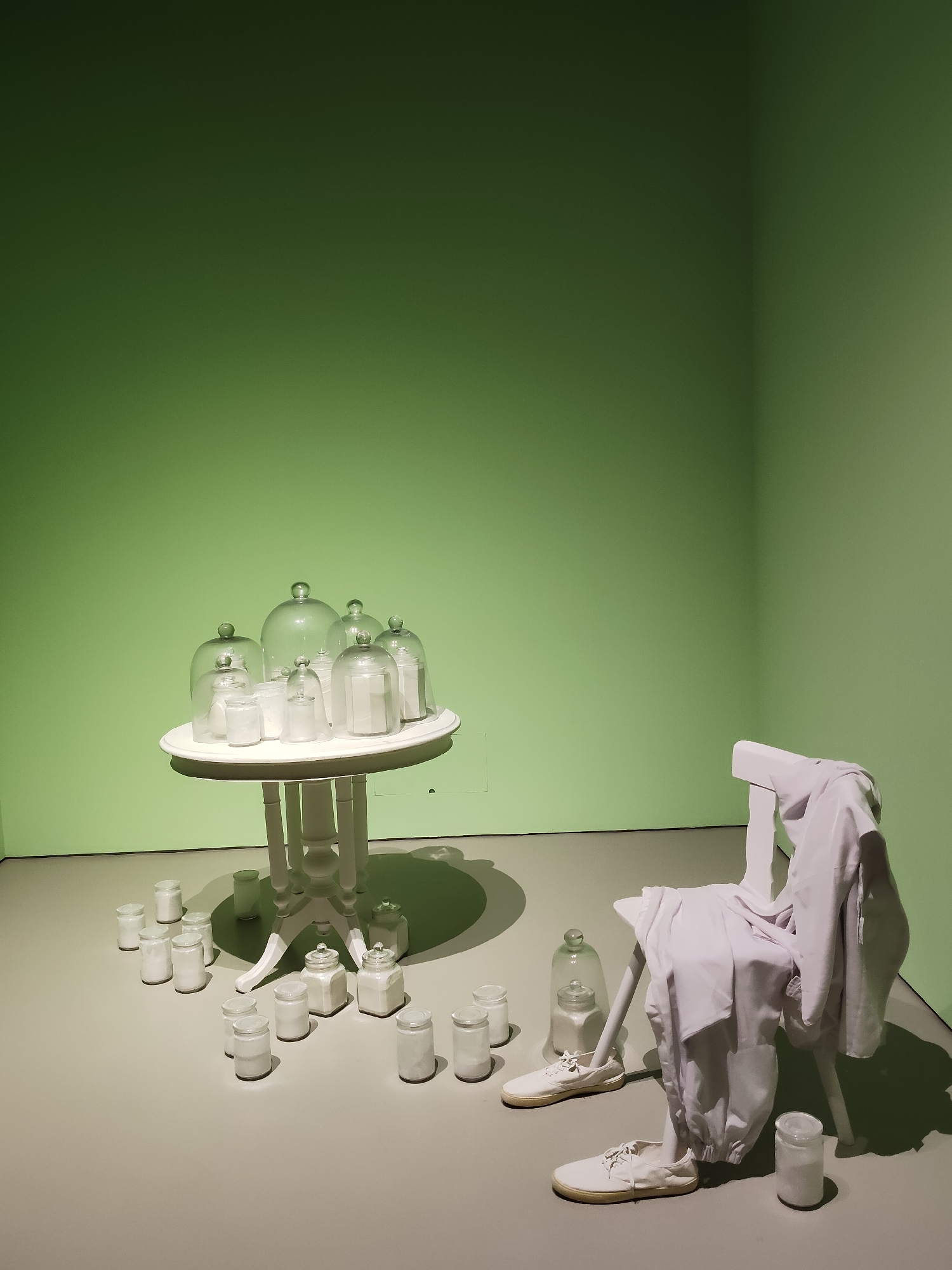 An art installation features a white, multi-legged table laden with glass cloches containing white powder, positioned in front of a pale green wall. Surrounding the table on the floor are numerous small white jars also filled with white powder. To the right, a white chair draped with white fabric is set on the floor, with a pair of white sneakers placed in front of it. The scene is lit from above, casting shadows on the floor. The installation appears to be in a gallery space, possibly in Singapore, as there are no distinct architectural features to pinpoint a specific location. There are no people present in the image.