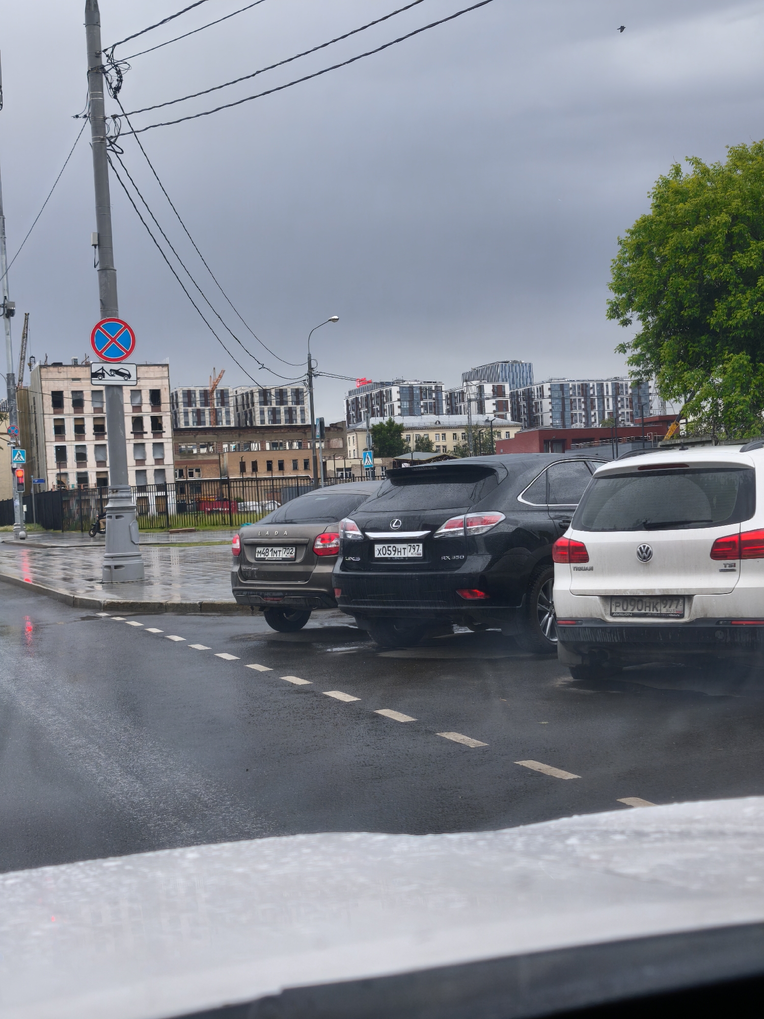 The image shows a street scene in Moscow, Russia, taken on a wet day. Three cars are parked close together along a road with white dotted lines, the view is from inside a vehicle. A brown Lada with license plate "M481MT 702", a black Lexus RX 450 with license plate "X059HT 797", and a white Volkswagen Tiguan with license plate "P090HK 777". Behind the cars are buildings, a traffic sign indicating no parking and towing, and a tree on the right. The sky is overcast.